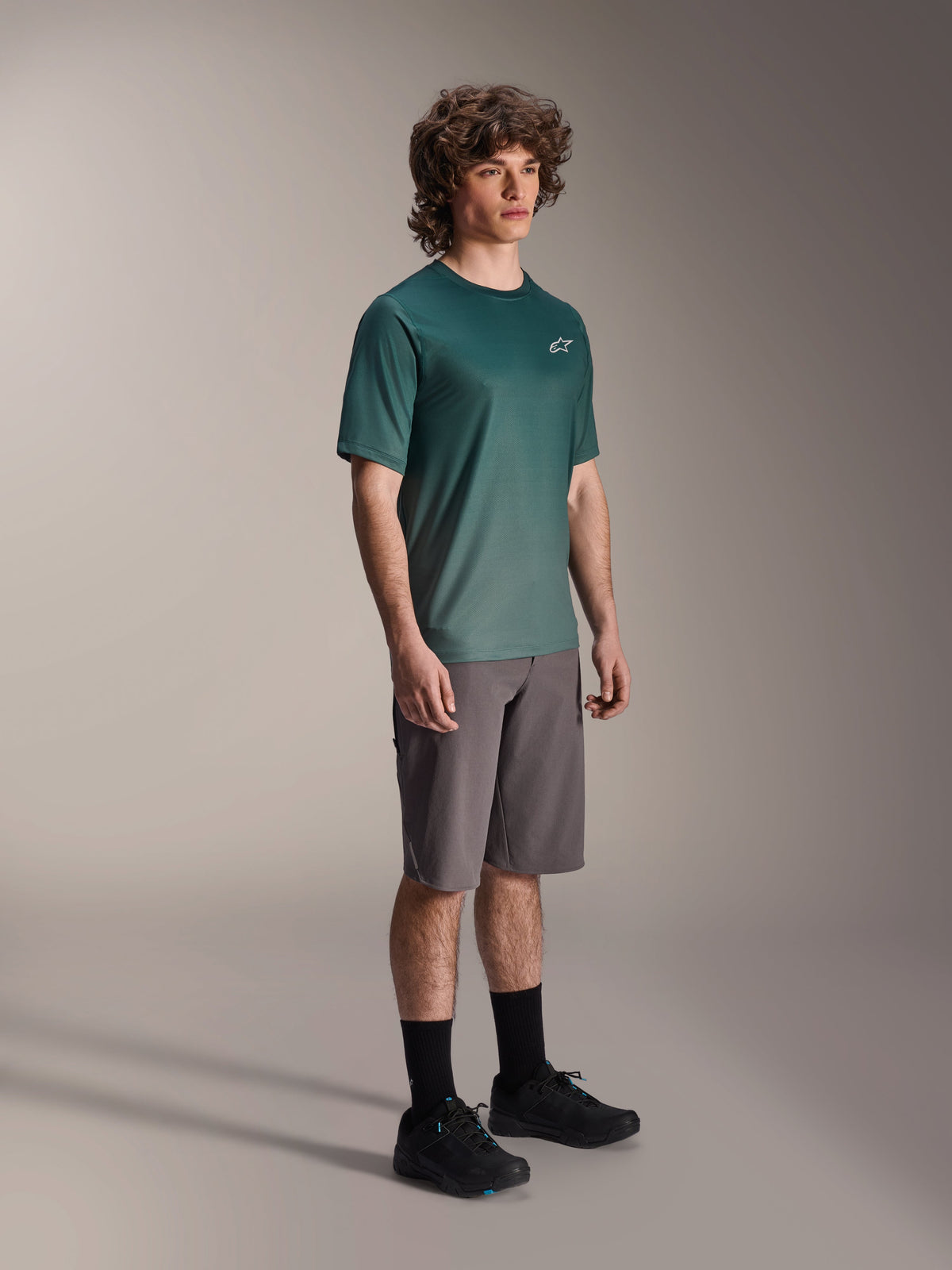 Alpinestars Maglia A-Dura Rise - Manica Corta in Silver Pine, maglia da mountain bike verde indossata da un modello con pantaloncini grigi e sneakers nere, abbigliamento casual per trail riding MTB con un piccolo logo bianco sul petto