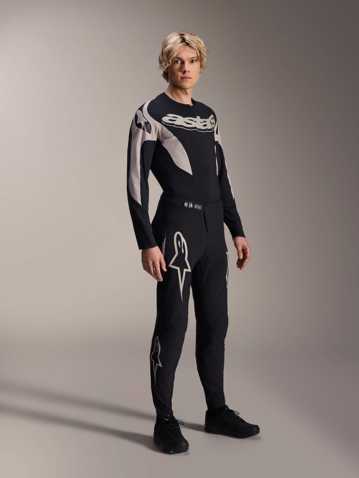 Modello che indossa i pantaloni Alpinestars A-Supra Melt e la maglia a maniche lunghe coordinata, nero con motivi curvi grigio chiaro, grandi loghi Astars bianchi sulle gambe e scritta sul petto, abbigliamento professionale da gara per mountain bike