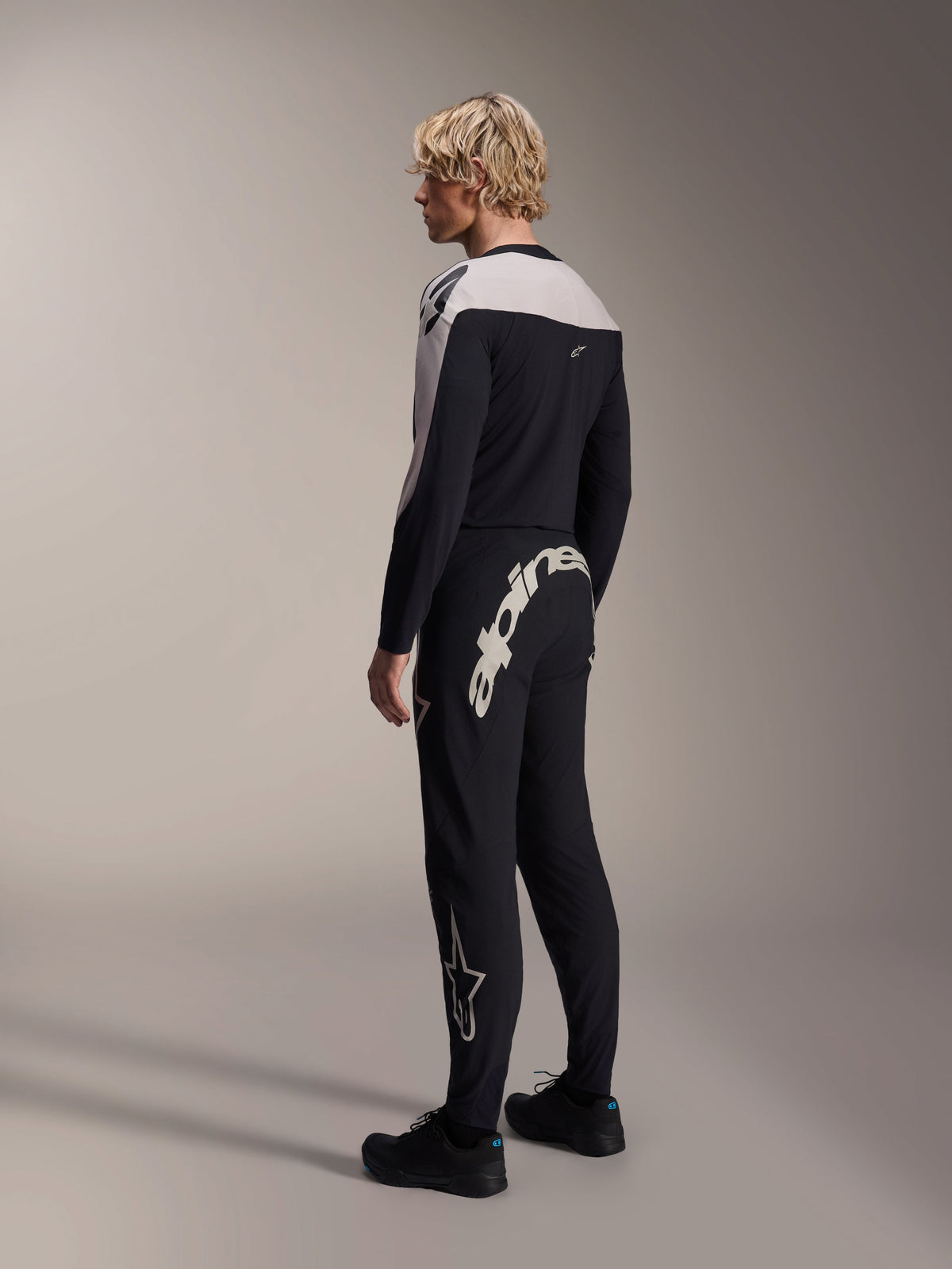 Alpinestars A-Supra Melt Pants, pantaloni da MTB, nero, caratterizzati da una grande scritta Alpinestars bianca sul carré posteriore e logo a stella sulla gamba, indossati da un modello con maglia a maniche lunghe coordinata e scarpe nere, vista posteriore laterale