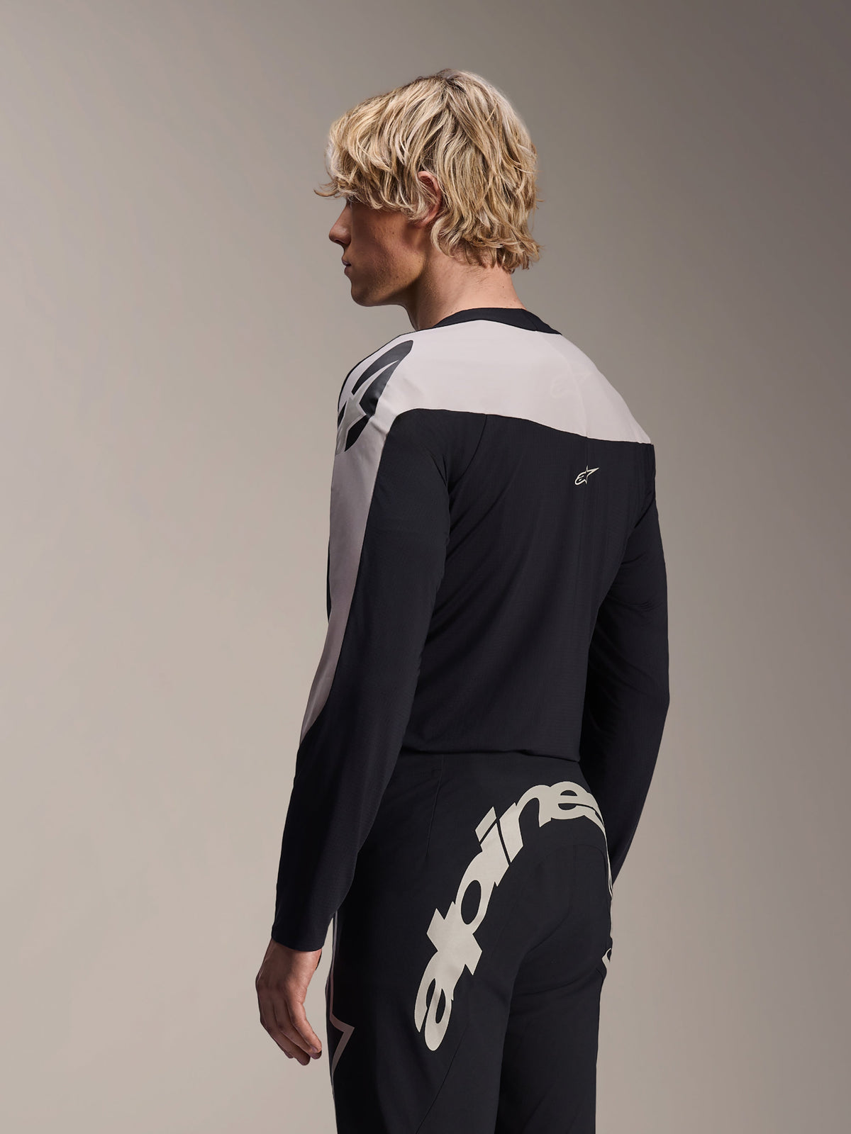 Modello uomo con maglia a maniche lunghe Alpinestars A-Supra Melt e pantaloni A-Supra Melt, completo da gara MTB nero e grigio chiaro, con grande logo Alpinestars bianco sul carré posteriore dei pantaloni