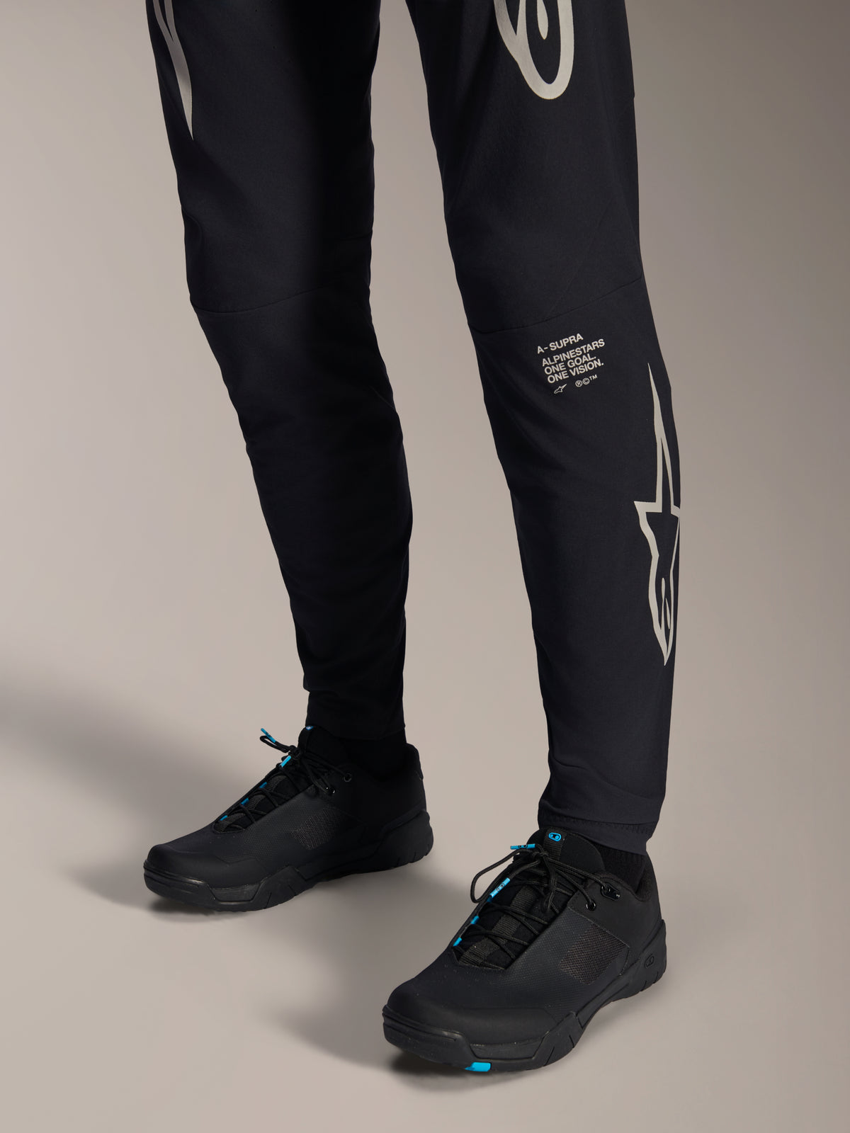 Pantaloni Alpinestars A-Supra Melt, pantaloni da mountain bike neri, con scritta e logo Alpinestars bianchi sulla gamba, indossati con scarpe da ciclismo nere con accenti blu, abbigliamento tecnico per fuoristrada