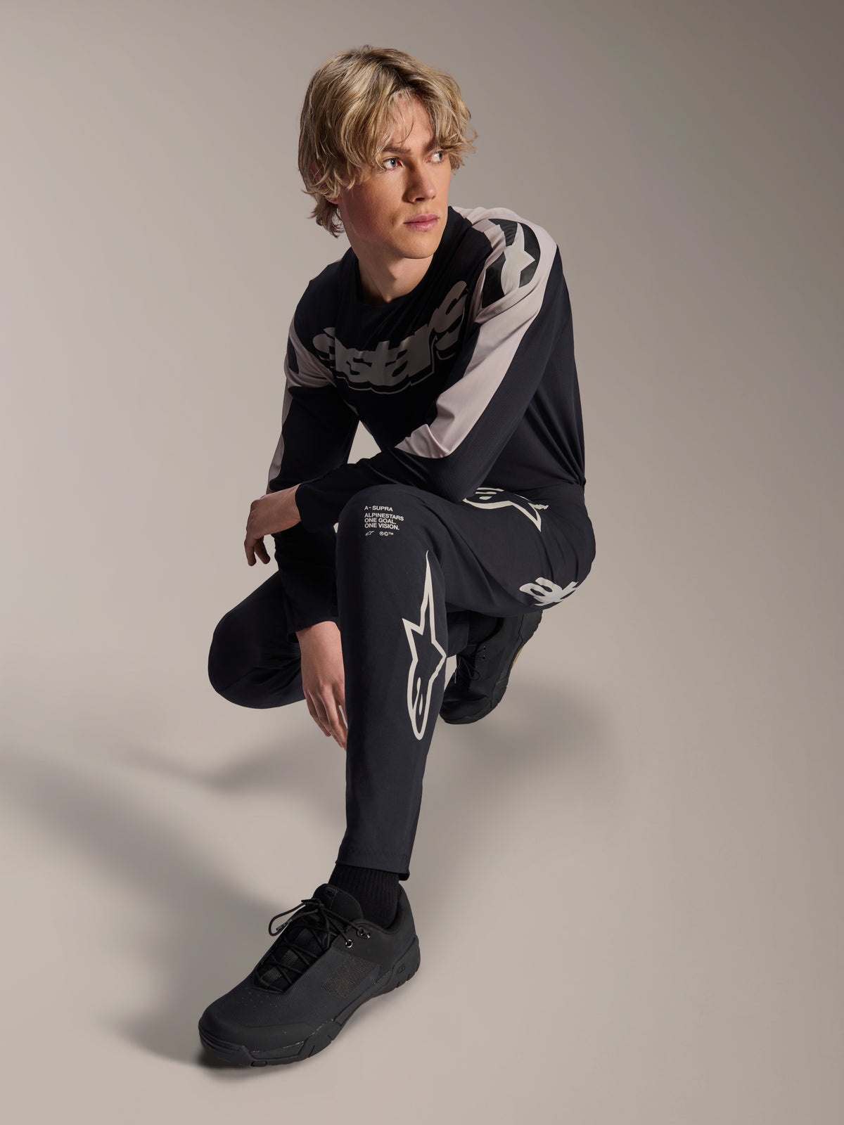 Pantaloni e maglia a maniche lunghe Alpinestars A-Supra Melt, nero e grigio, caratterizzati da audace branding bianco e loghi a stella, indossati da un modello maschile in posa accovacciata, completo coordinato da gara MTB.