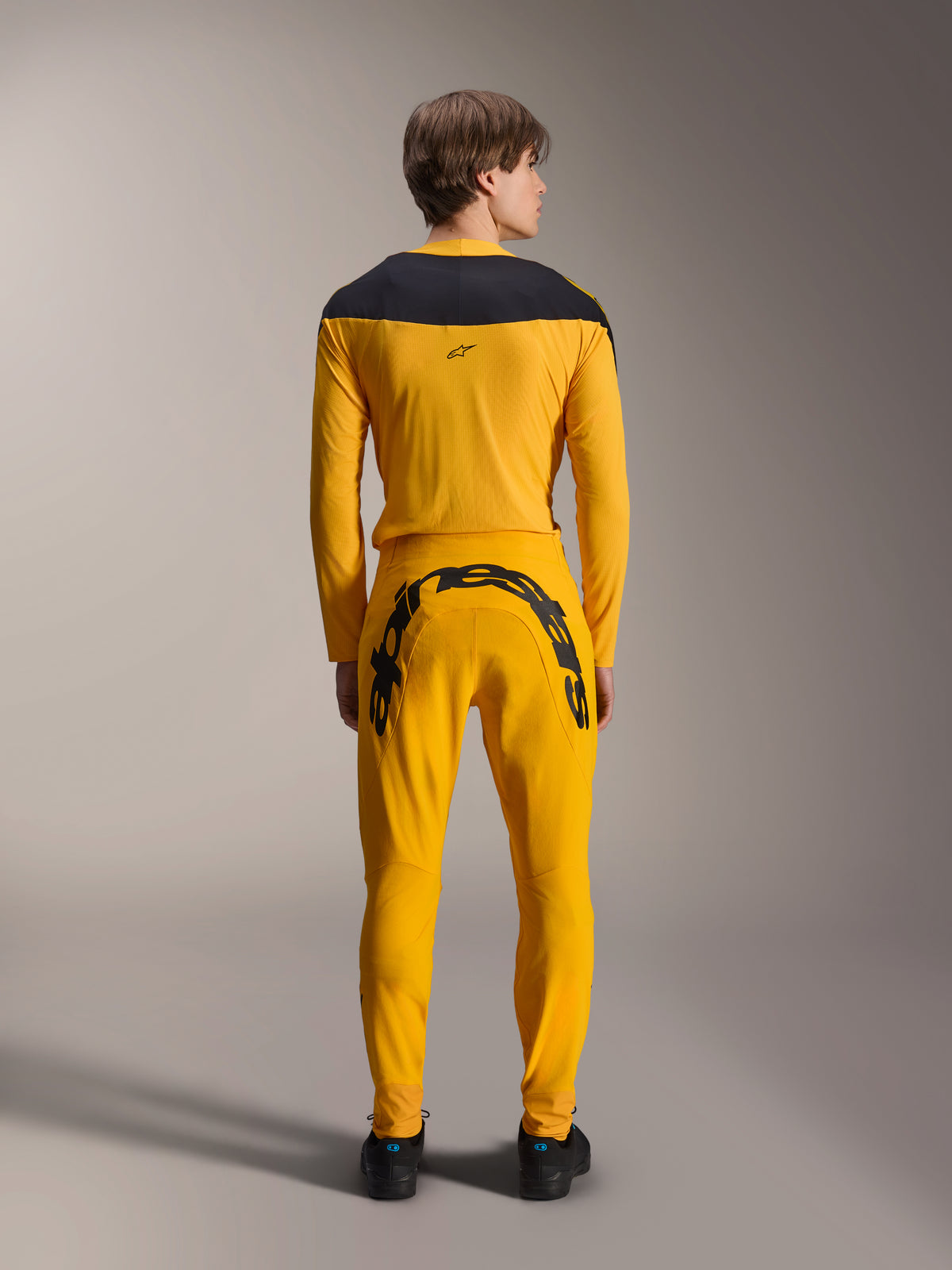 Vista posteriore di un modello che indossa il set completo Alpinestars Limited Edition A-Supra Melt Gold, composto da una maglia a maniche lunghe giallo scuro con carré nero sulle spalle e pantaloni coordinati con grande scritta Alpinestars nera sul carré posteriore, abbinato a scarpe nere.