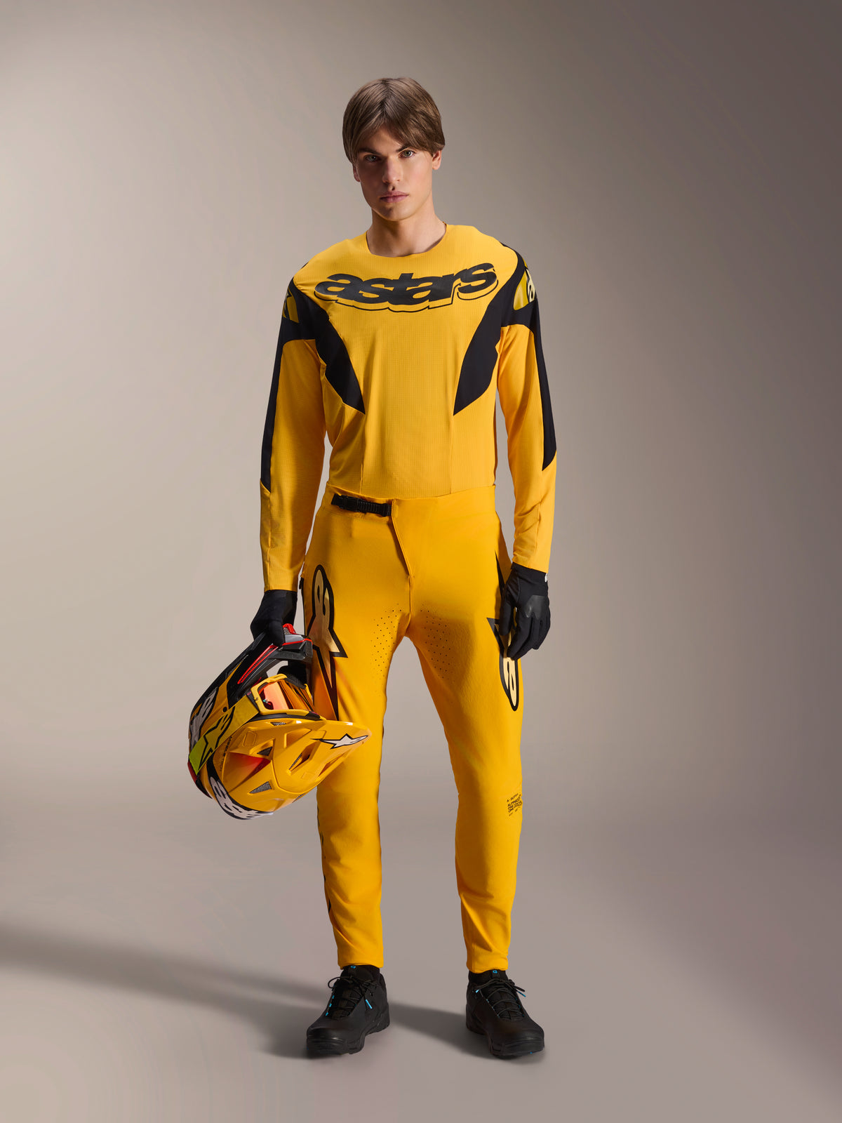 Modello con Kit MTB Alpinestars Limited Edition A-Supra Melt Gold, maglia gialla a maniche lunghe con logo 'astars' nero, pantaloni gialli coordinati con dettagli neri, con in mano un casco da gara giallo e nero, guanti e scarpe neri, abbigliamento tecnico per mountain bike