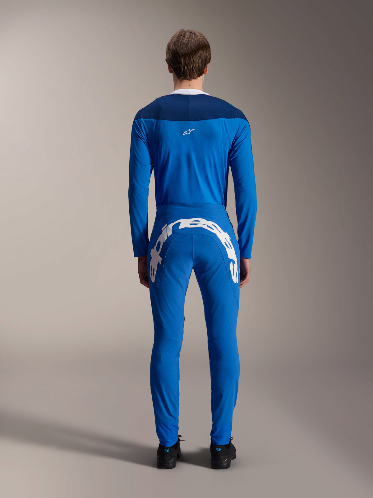 Modello maschile visto di spalle che indossa la maglia Alpinestars A-Supra Melt a maniche lunghe in blu, pannello sulle spalle blu scuro con piccolo logo a stella bianco, pantaloni MTB coordinati con grande marchio Alpinestars bianco sul retro, outfit tecnico per mountain bike.