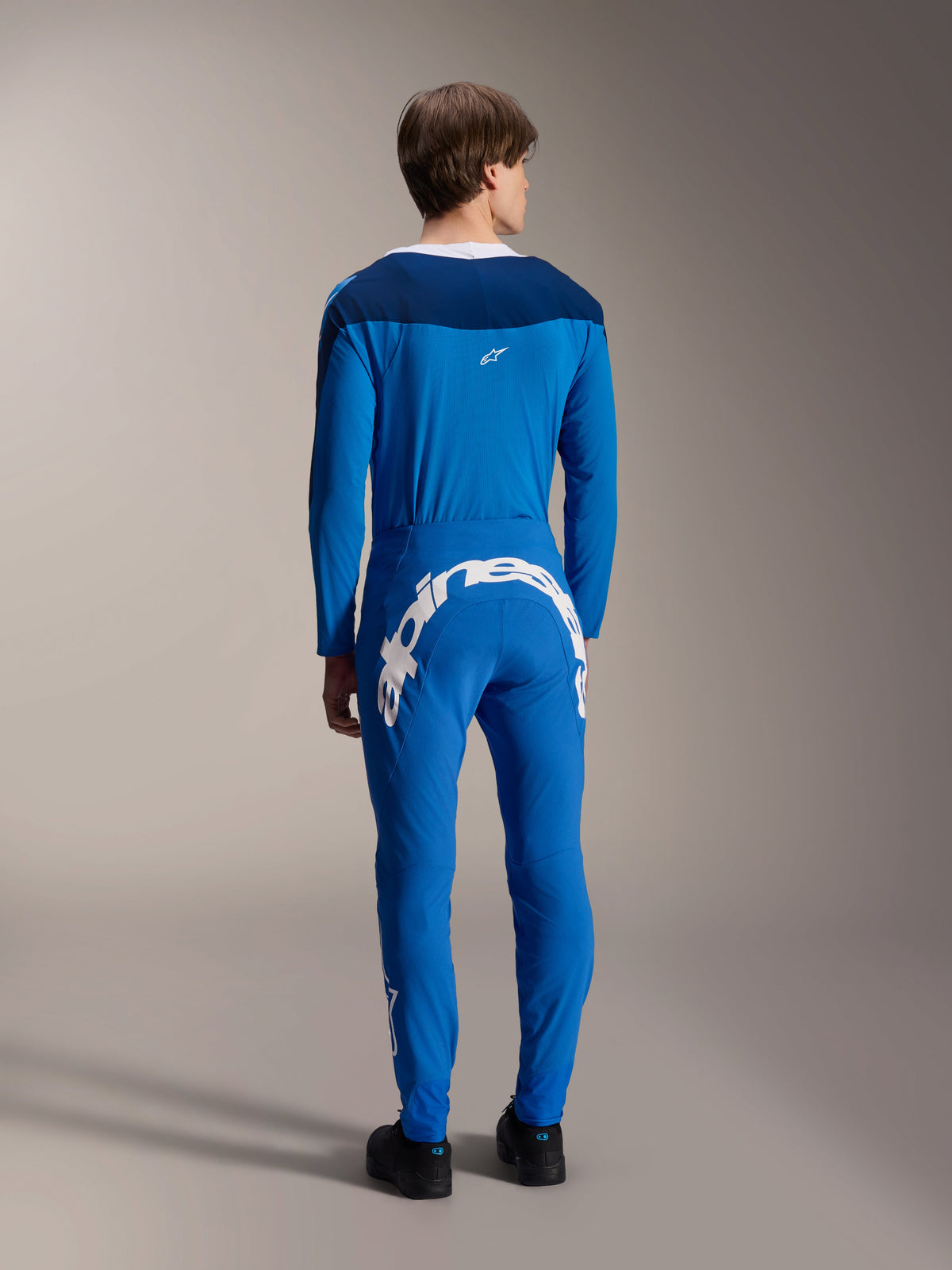 Modello maschile visto dal retro che indossa la maglia a maniche lunghe Alpinestars A-Supra Melt in blu, abbinata a pantaloni tecnici blu coordinati con un grande logo Alpinestars bianco sul retro, kit completo da gara MTB.