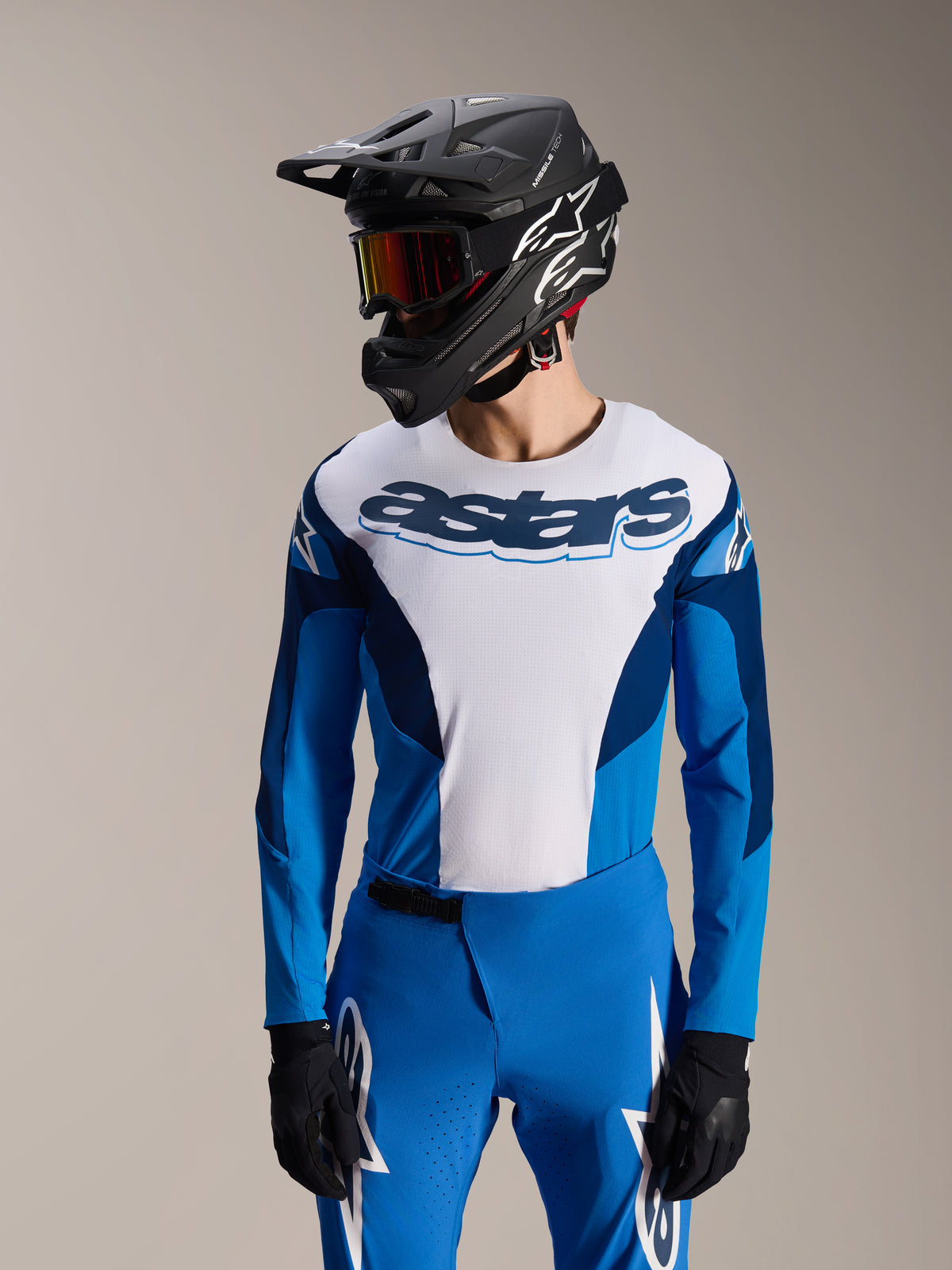 Modello che indossa la maglia Alpinestars A-Supra Melt a maniche lunghe in blu e bianco, con scritta 'astars' sul petto, abbinata a pantaloni da MTB blu coordinati, casco integrale nero e maschera, completo da gara per mountain bike.
