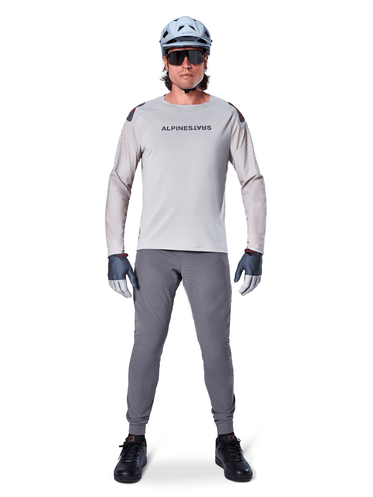 Maglia Alpinestars A-Aria Polartec Switch, a maniche lunghe, grigio chiaro, indossata da un ciclista di mountain bike con pantaloni MTB grigio scuro, casco azzurro chiaro e occhiali da sole, con marchio sul petto e ventilazione traforata al laser sulle maniche, outfit completo da riding off-road.
