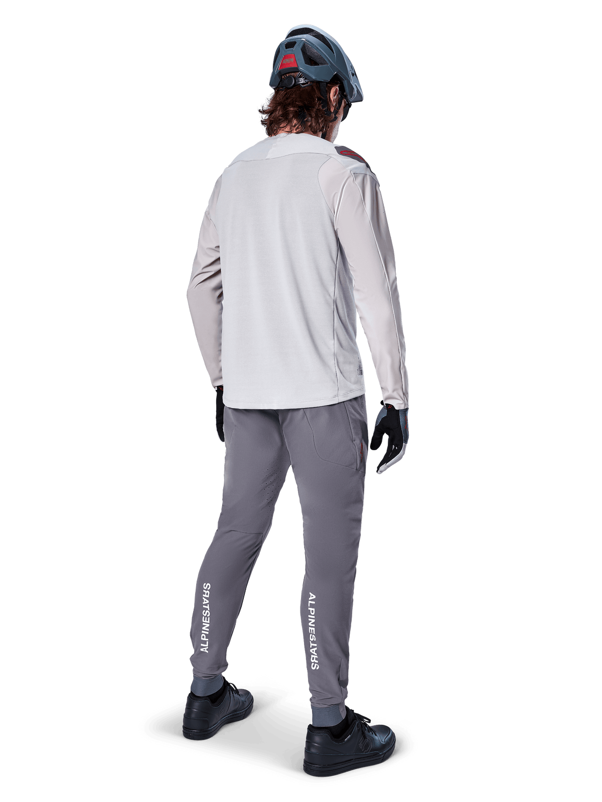 Vista posteriore di un ciclista che indossa la maglia a maniche lunghe Alpinestars A-Aria Polartec Switch in grigio chiaro, abbinata a pantaloni da mountain bike grigio scuro con marchio Alpinestars bianco sui polpacci, guanti neri e un casco grigio