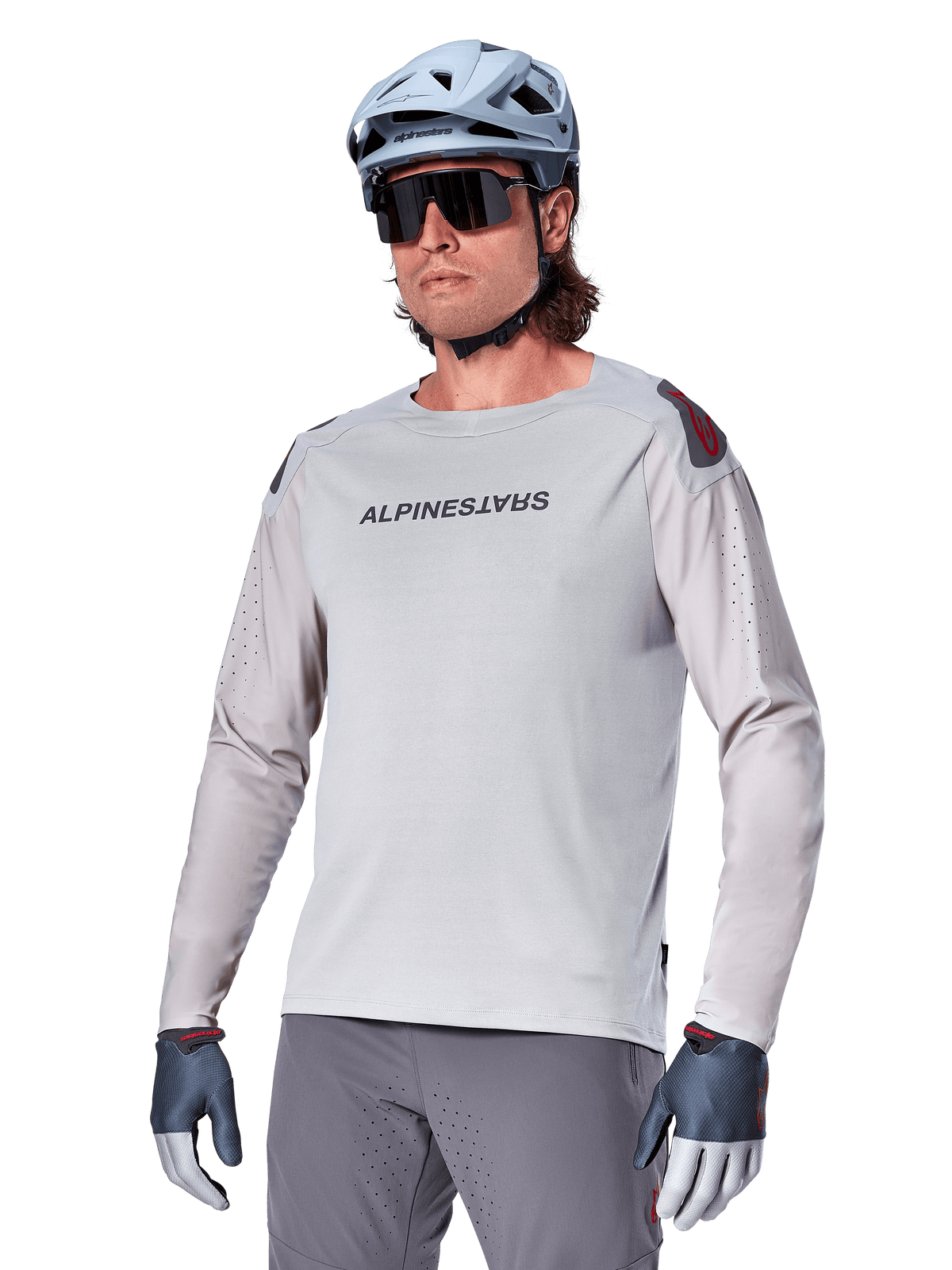 Alpinestars A-Aria Polartec Switch Jersey a maniche lunghe, maglia da ciclismo grigio chiaro con logo nero sul petto e dettaglio rosso sulla spalla, maniche perforate al laser per la ventilazione, indossata con casco azzurro, occhiali da sole e pantaloni grigi, abbigliamento tecnico da mountain bike per trail