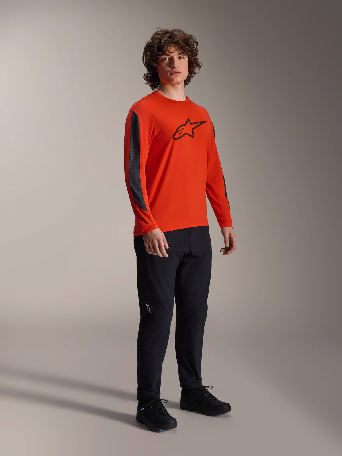Modello che indossa la maglia Alpinestars A-Dura Insert Astar a maniche lunghe, Spicy Orange con logo Astar nero sul petto e inserti grigio scuro sulle maniche, indossata con pantaloni neri, abbigliamento professionale per ciclismo MTB