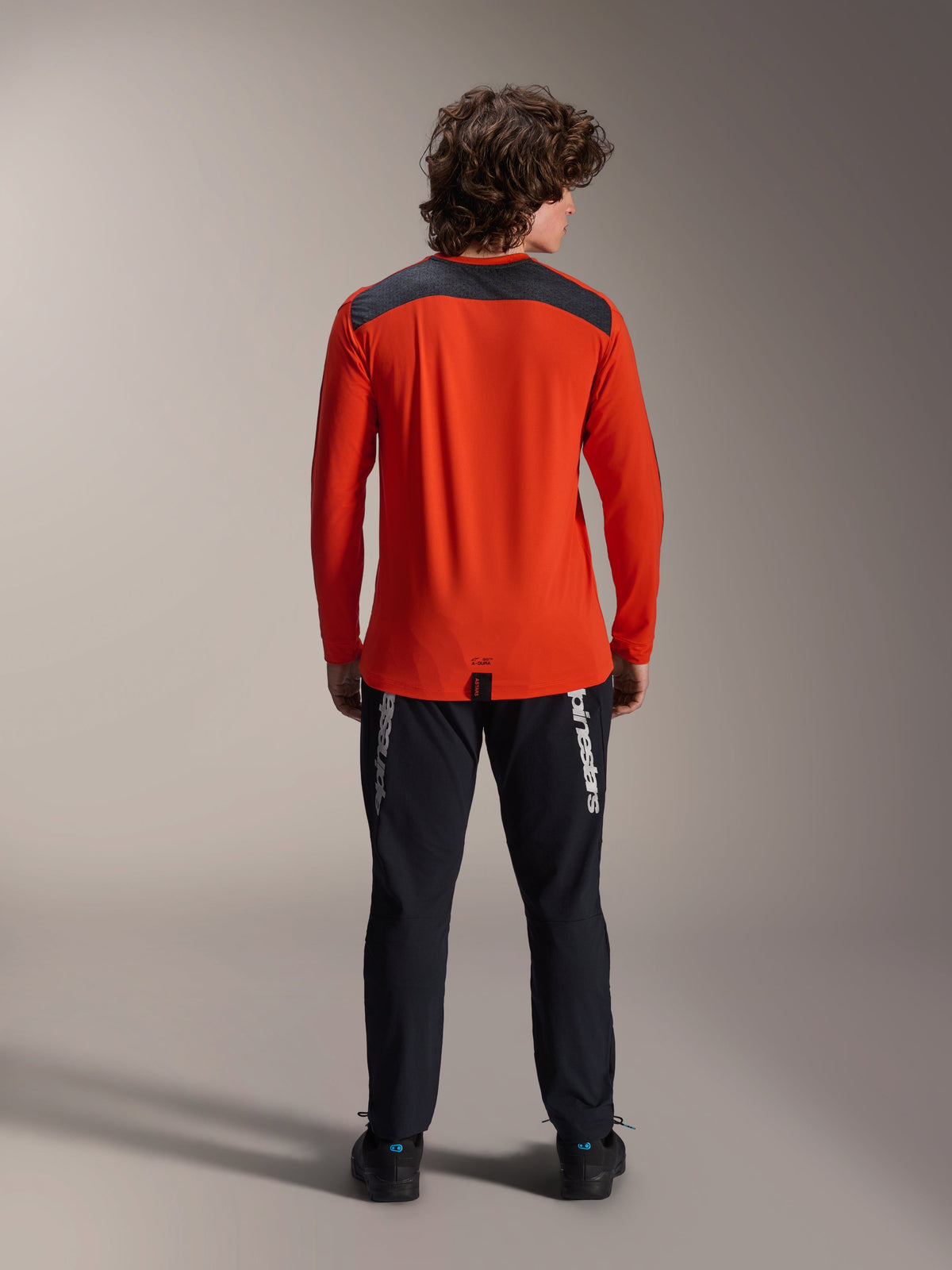 Maglia Alpinestars A-Dura Insert Astar a maniche lunghe, Spicy Orange, vista posteriore con pannello sulle spalle grigio antracite, indossata con pantaloni Alpinestars neri e scarpe nere, scatto professionale in studio per abbigliamento tecnico da MTB