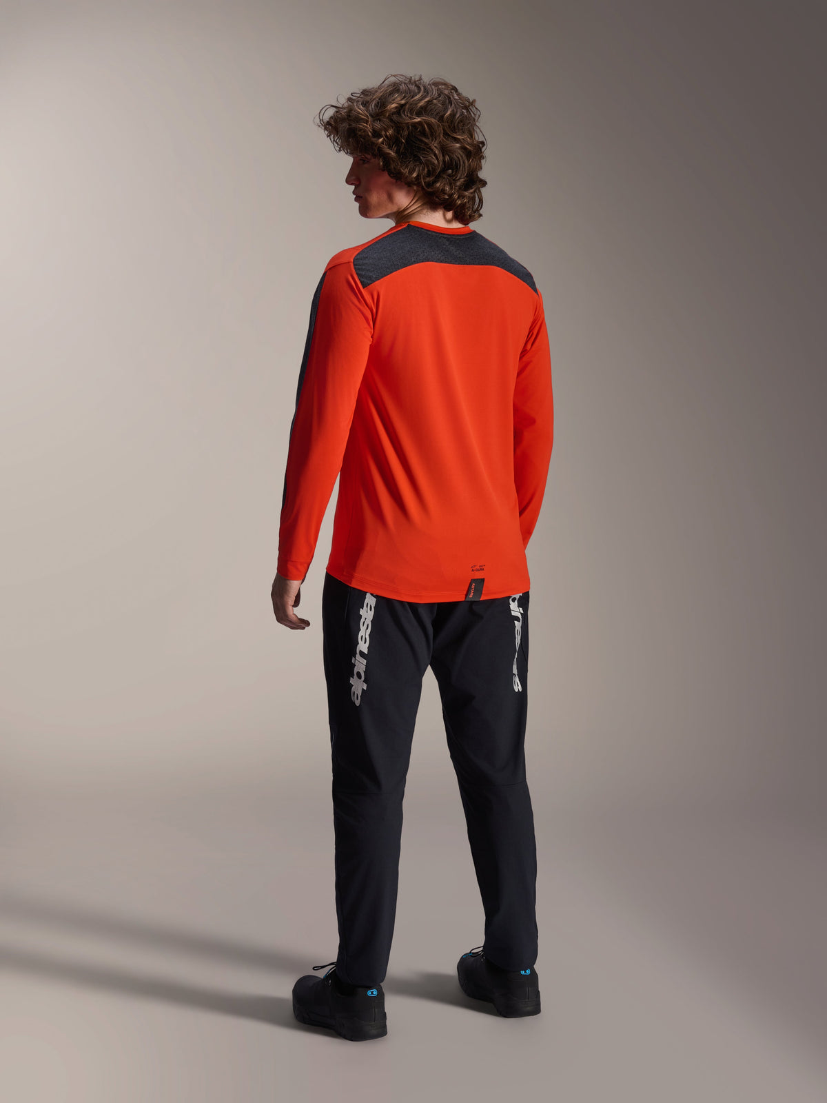 Vista posteriore di un modello che indossa la maglia Alpinestars A-Dura Insert Astar in Spicy Orange, maglia da MTB a maniche lunghe con pannello sulle spalle grigio scuro, abbinata a pantaloni neri con marchio, abbigliamento da ciclismo off-road.