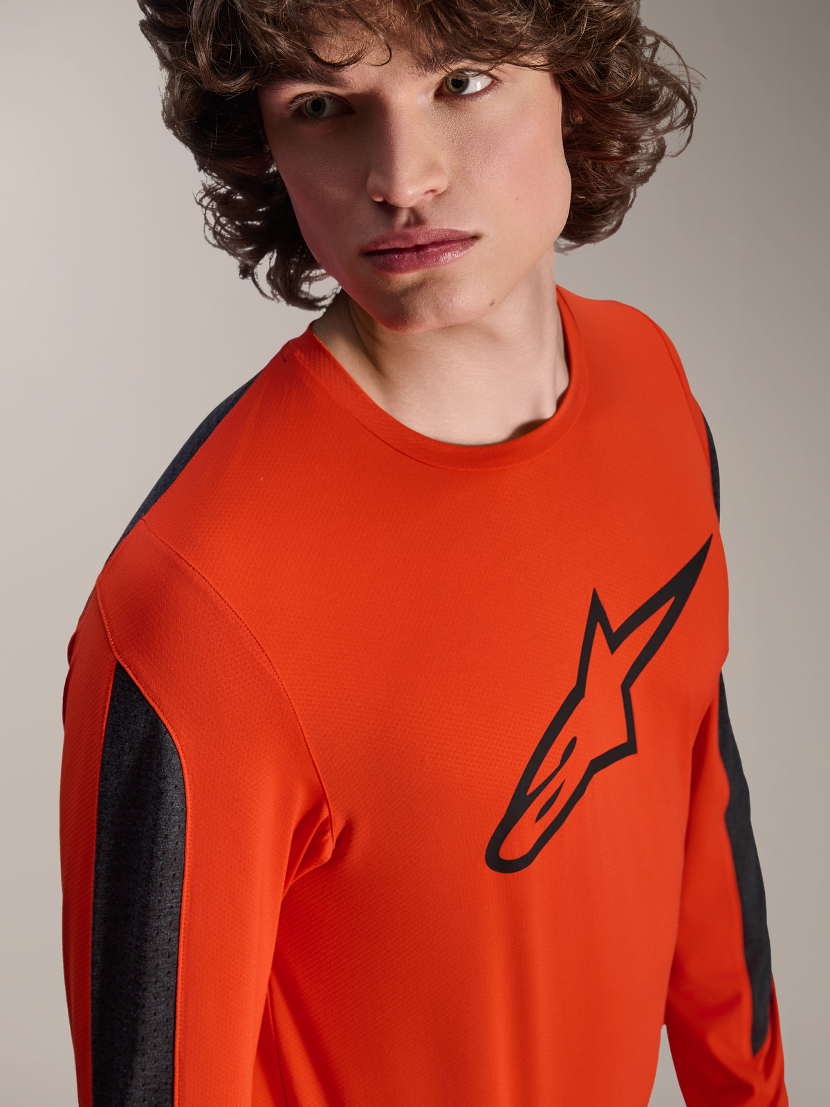 Maglia Alpinestars A-Dura Insert Astar a maniche lunghe, maglia da MTB Spicy Orange con grande logo Astar nero sul petto, pannelli sulle maniche grigio scuro, indossata da un modello in un primo piano in studio, tessuto tecnico testurizzato