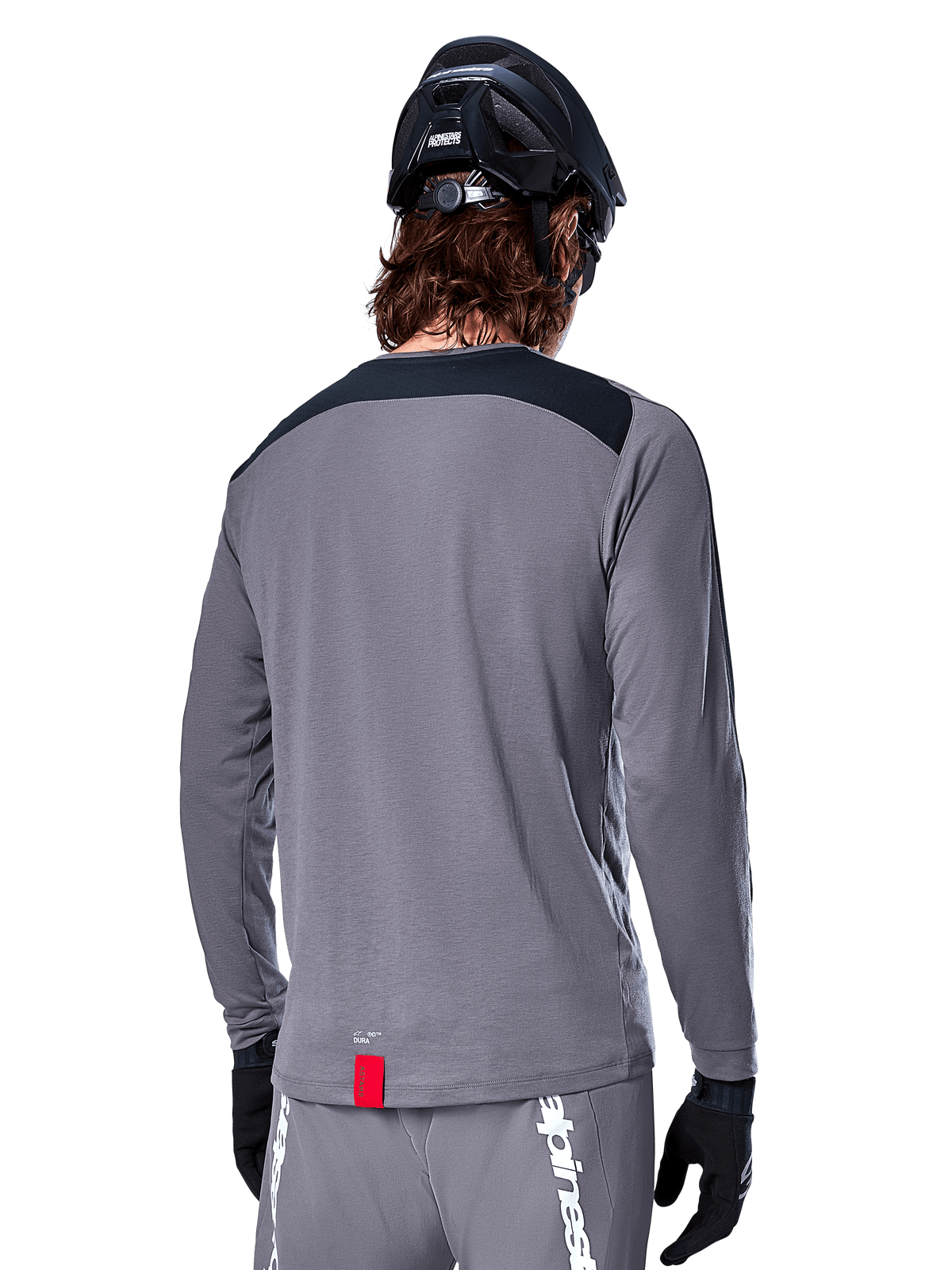 Alpinestars Maglia A-Dura Dri Astar a maniche lunghe, maglia da MTB grigio scuro vista dal retro, con pannello nero sulle spalle e piccola etichetta rossa del brand, indossata con casco e guanti da mountain bike neri