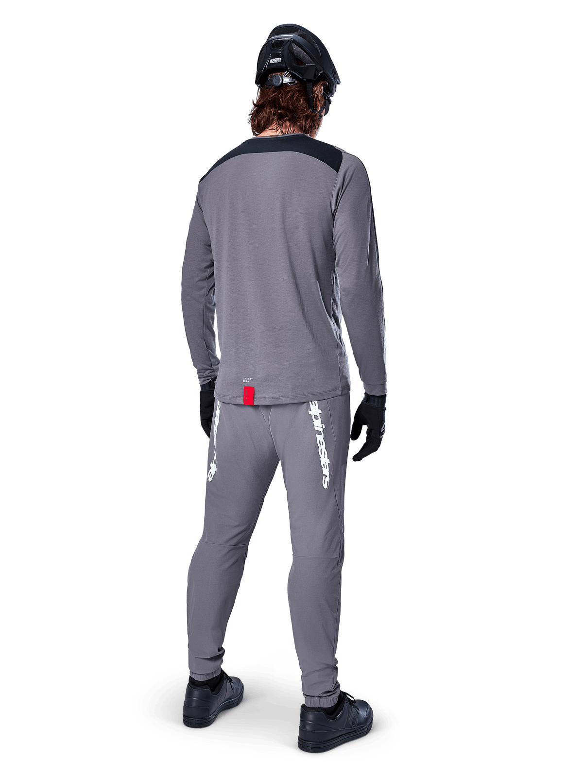 Maglia Alpinestars A-Dura Dri Astar - Maniche Lunghe, grigio scuro, vista posteriore del ciclista con pantaloni abbinati e casco nero, presenta pannelli neri sulle spalle e una piccola etichetta rossa sull'orlo, abbigliamento tecnico da ciclismo fuoristrada