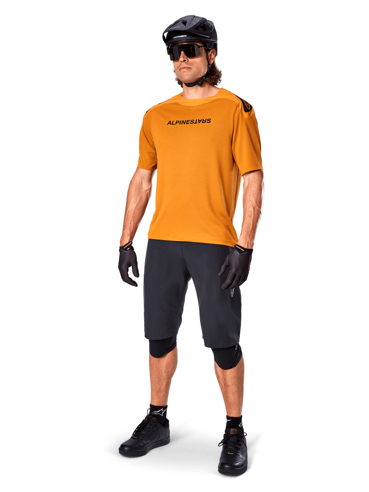 Ciclista che indossa la maglia Alpinestars A-Aria Polartec Switch in Dark Gold, maglia da MTB a maniche corte, abbinata a pantaloncini da ciclismo neri, ginocchiere, casco nero e guanti, abbigliamento professionale da mountain bike per trail riding