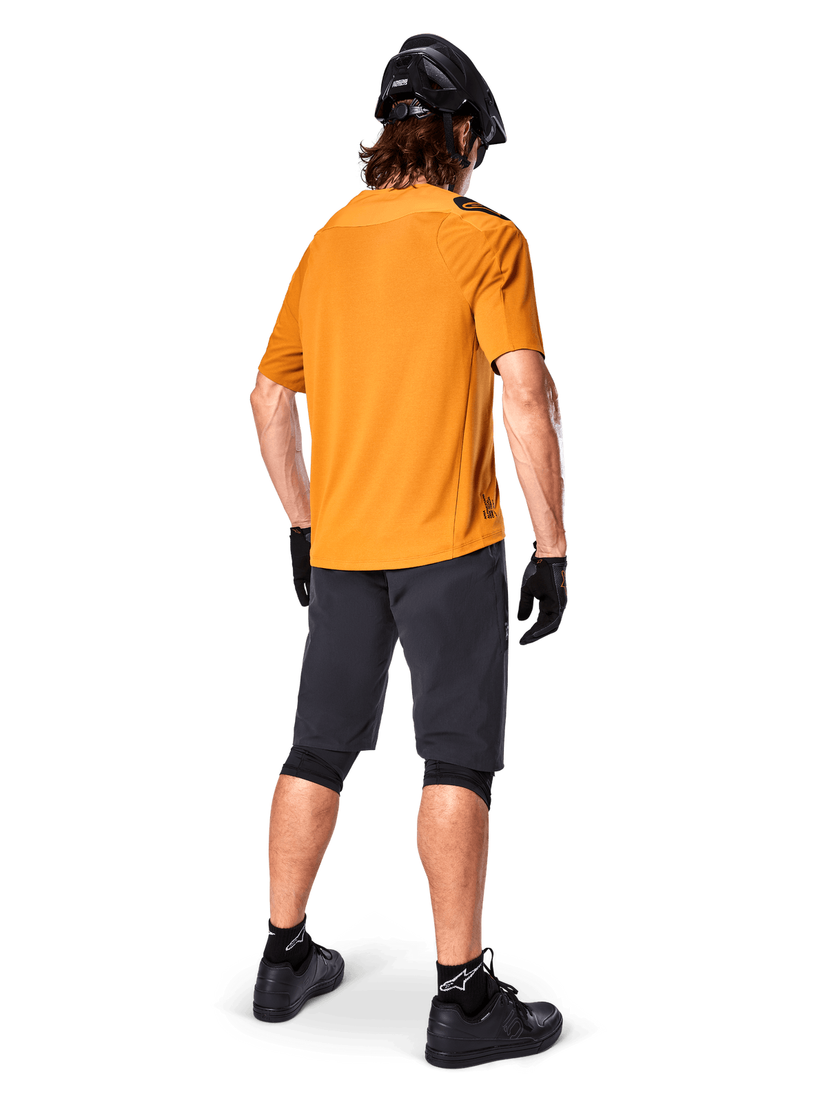 Alpinestars Maglia A-Aria Polartec Switch, maglia MTB a maniche corte, oro scuro, vista posteriore del modello con pantaloncini da mountain bike grigio scuro, casco nero e guanti, abbigliamento tecnico da ciclismo per percorsi fuoristrada
