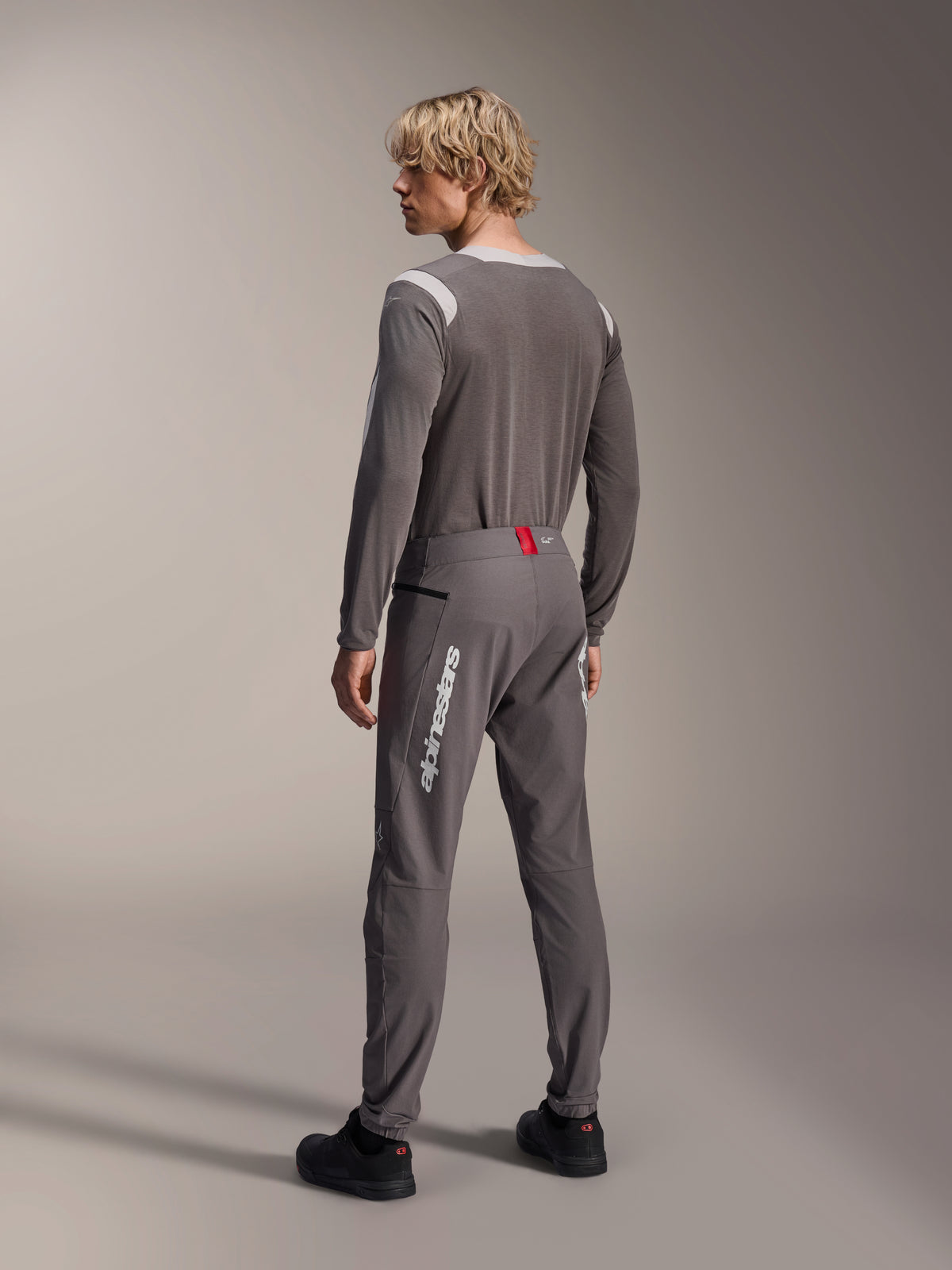Modello visto da dietro con abbigliamento Alpinestars MTB, maglia a maniche lunghe A-Dura Dri Wool grigia, pantaloni A-Dura Elite grigio scuro con logo verticale bianco e dettaglio rosso in vita, completo coordinato da mountain bike