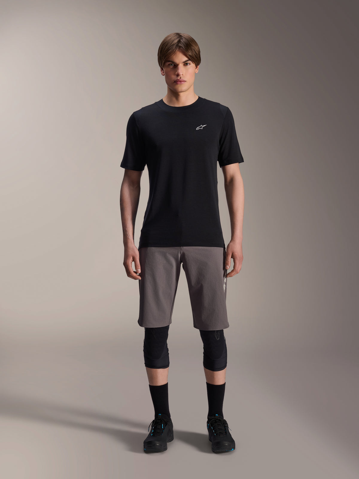 Modello con pantaloncini Alpinestars A-Dura Elite in grigio scuro e maglia a maniche corte A-Dura Dri-Wool in nero, abbinati a ginocchiere nere e scarpe da ciclismo, abbigliamento tecnico da mountain bike per prestazioni off-road