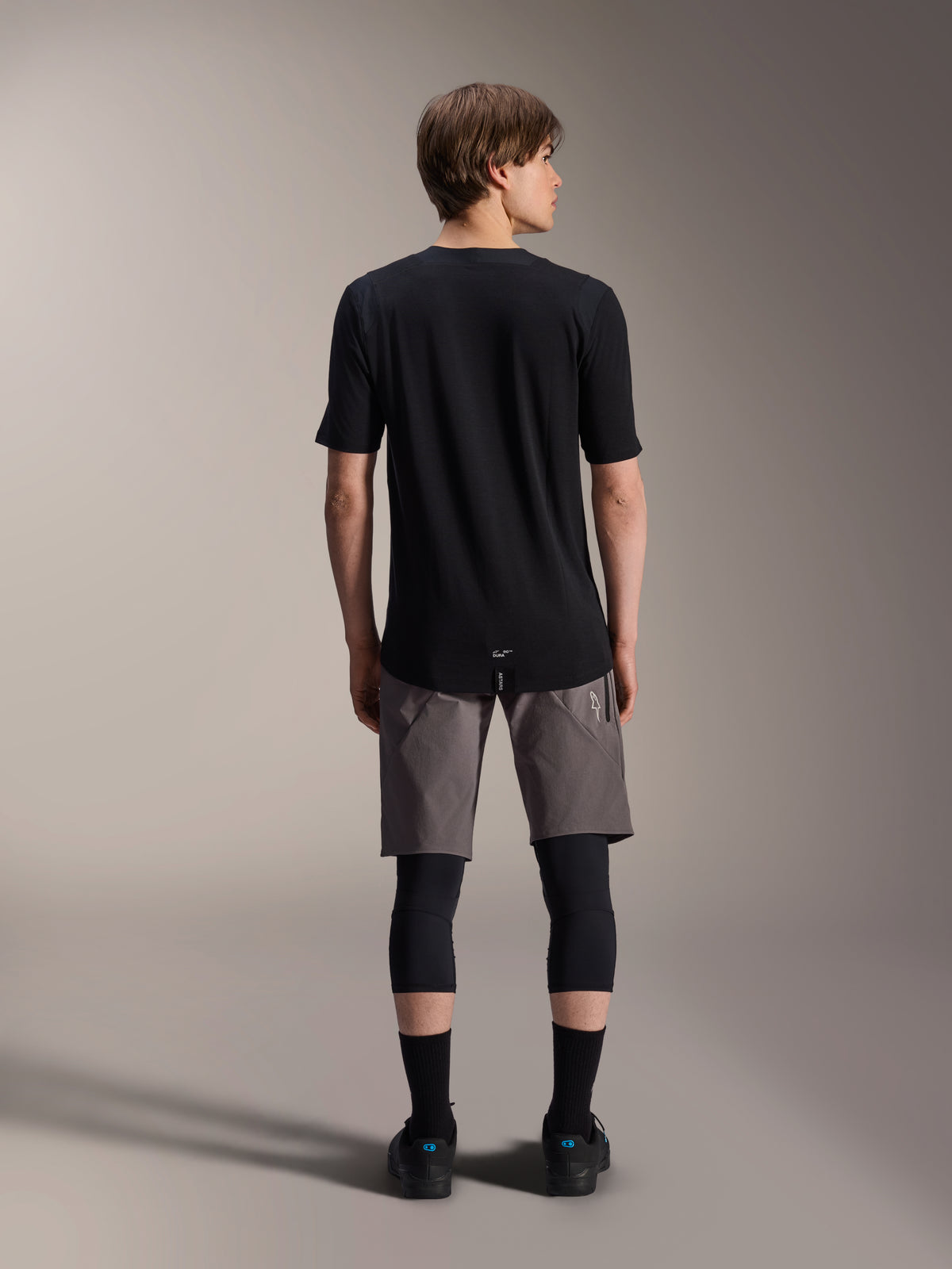 Alpinestars A-Dura Elite Shorts, pantaloncini da mountain bike grigio scuro, vista posteriore, indossati con maglia nera a maniche corte, ginocchiere e calze nere, abbigliamento tecnico da ciclismo per il fuoristrada