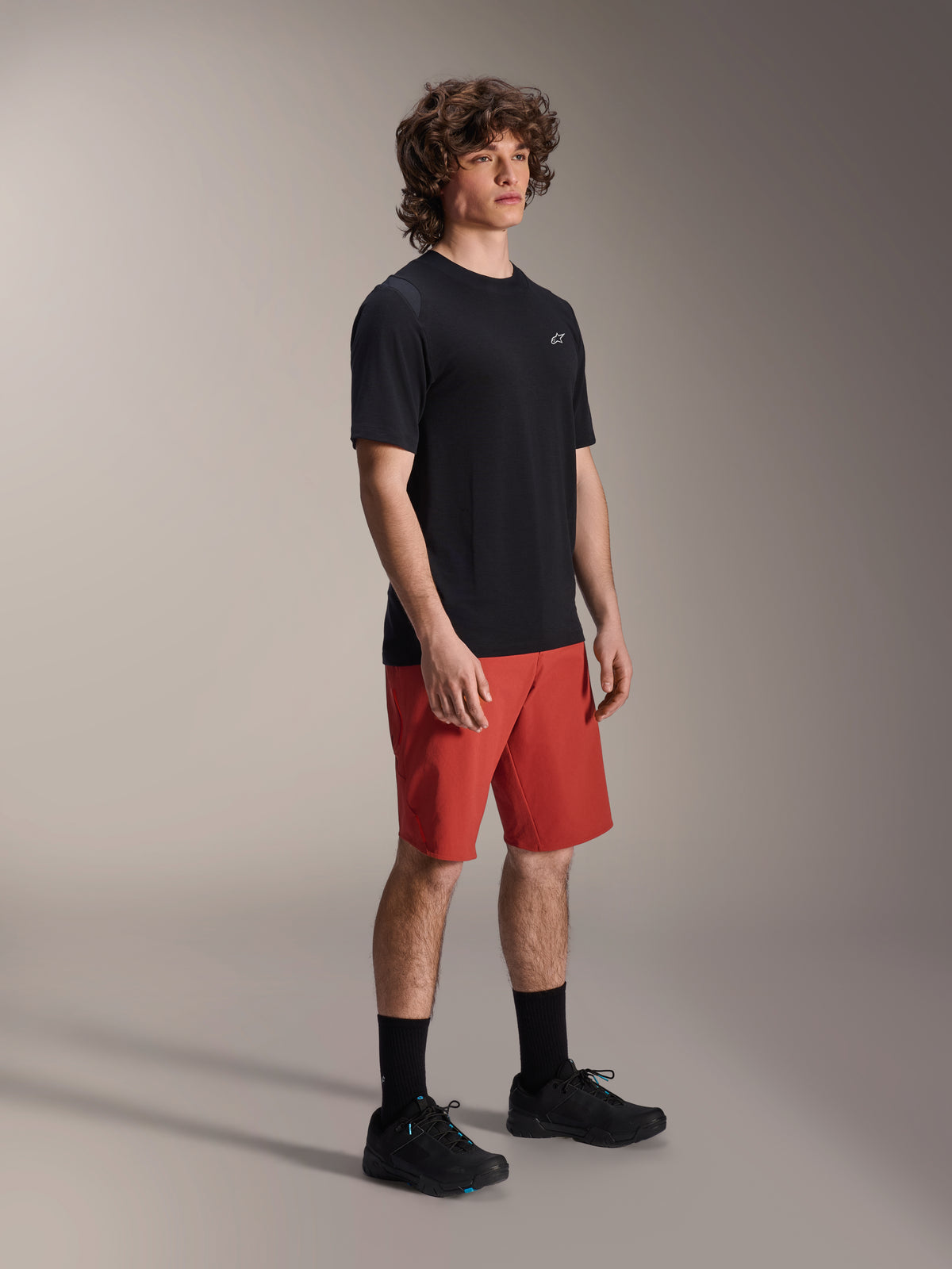 Modello uomo che indossa i pantaloncini Alpinestars A-Dura Elite in Dark Orange e la maglia a maniche corte A-Dura Dri Wool in nero, abbigliamento tecnico per mountain bike, abbinato a calze e scarpe nere, design sportivo per il ciclismo fuoristrada.
