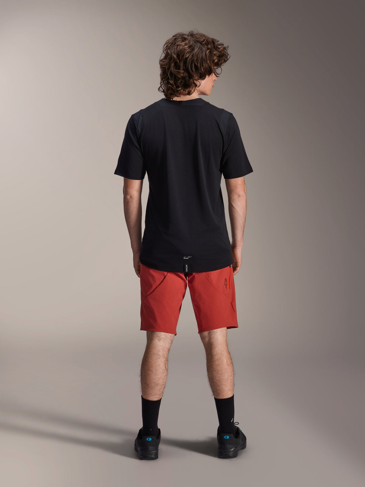 Modello che indossa i pantaloncini Alpinestars A-Dura Elite in arancione scuro e una maglia a maniche corte A-Dura Dri Wool nera, visto di spalle, abbinati a calze nere e scarpe da ciclismo, abbigliamento professionale da mountain bike