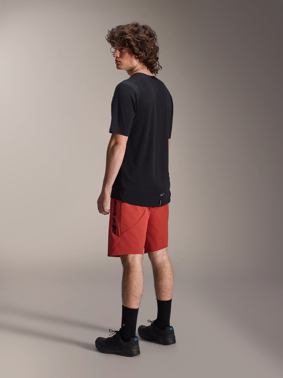 Alpinestars A-Dura Elite Shorts, pantaloncini da mountain bike, Dark Orange, indossati con una maglia nera a maniche corte e scarpe da mountain bike nere, vista laterale posteriore del modello che mette in risalto la vestibilità performante e il marchio