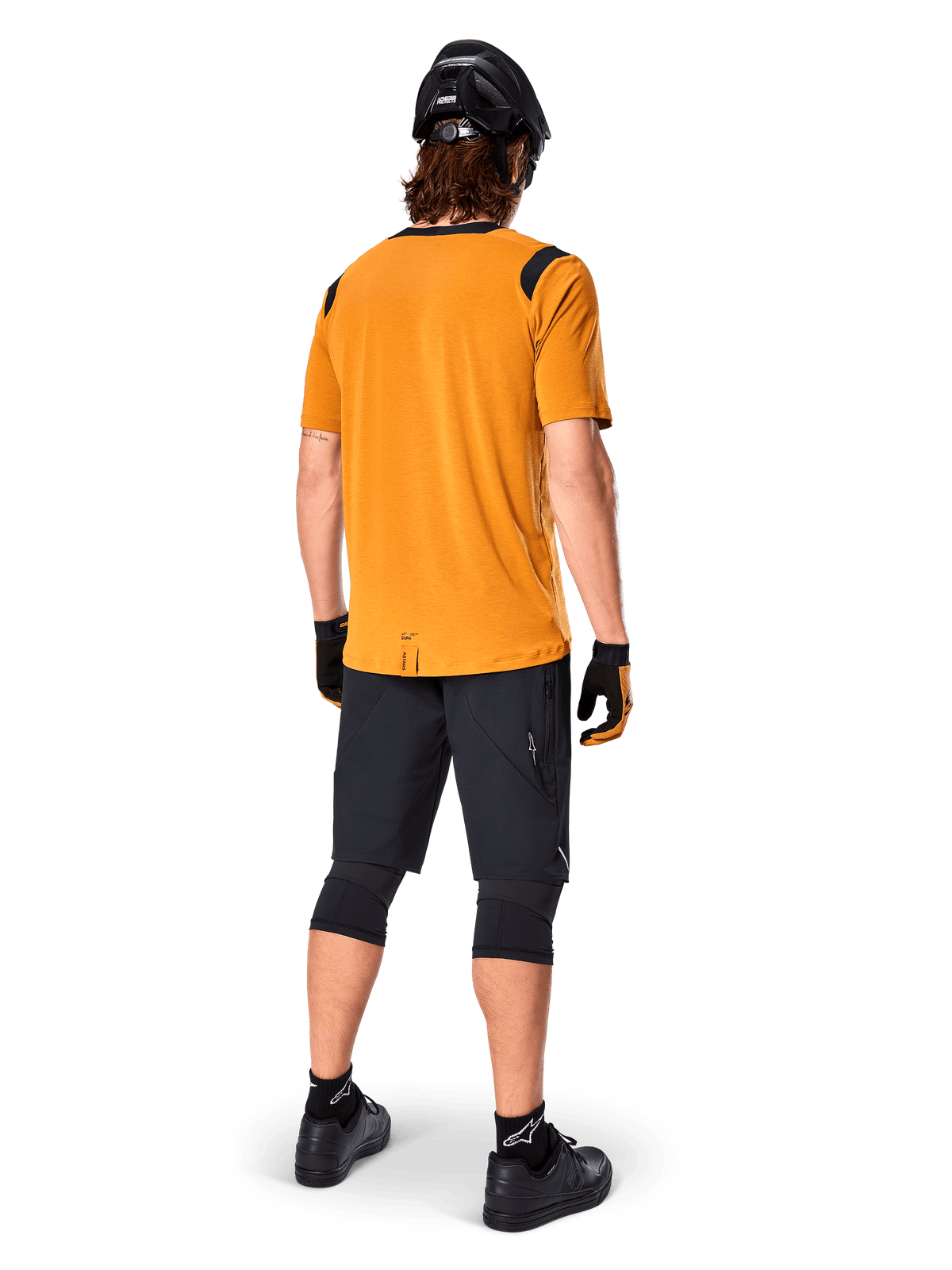 Alpinestars Maglia A-Dura Dri Wool a maniche corte, maglia da ciclismo, Dark Gold, vista posteriore di un mountain biker che indossa una maglia giallo senape con inserti in rete nera sulle spalle, abbinata a pantaloncini neri, ginocchiere, casco nero e guanti, abbigliamento tecnico da ciclismo fuoristrada
