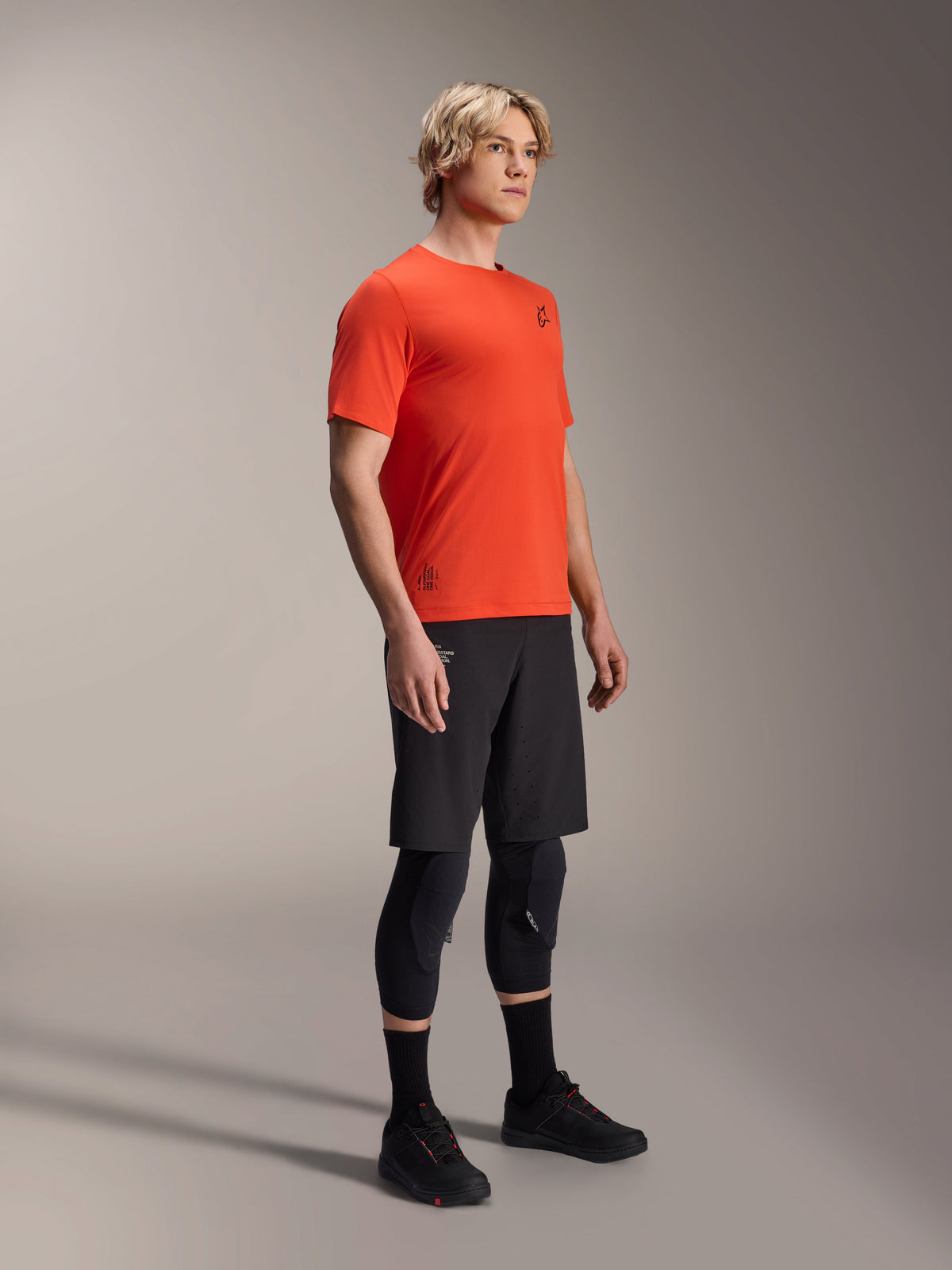 Maglia a maniche corte Alpinestars A-Aria Astar in Spicy Orange, indossata da un modello con pantaloncini MTB neri e ginocchiere, abbigliamento da mountain bike leggero e traspirante, arancione vivace con branding nero minimale