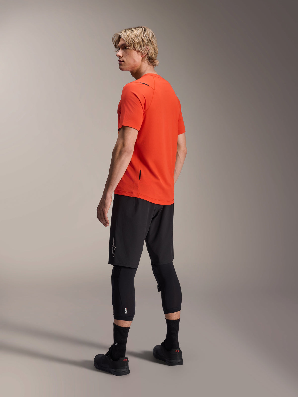 Maglia a maniche corte Alpinestars A-Aria Astar, Spicy Orange, vista di tre quarti posteriore sul modello che guarda oltre la spalla, abbinata a pantaloncini da MTB neri e ginocchiere, abbigliamento tecnico da mountain bike