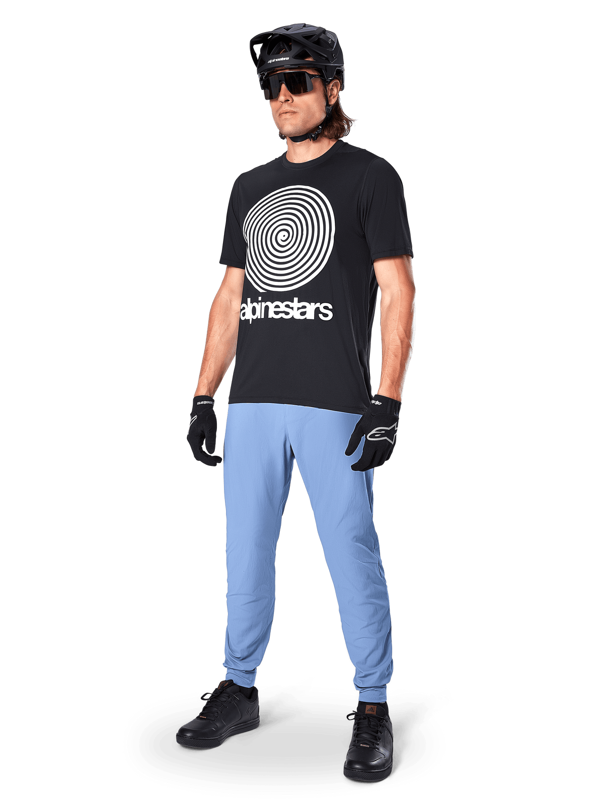 Maglia a maniche corte Alpinestars A-Dura Oscar, maglia da MTB nera con grande grafica a spirale heritage bianca, indossata da un rider con casco nero, occhiali da sole, guanti neri e pantaloni azzurri, abbigliamento tecnico da mountain bike