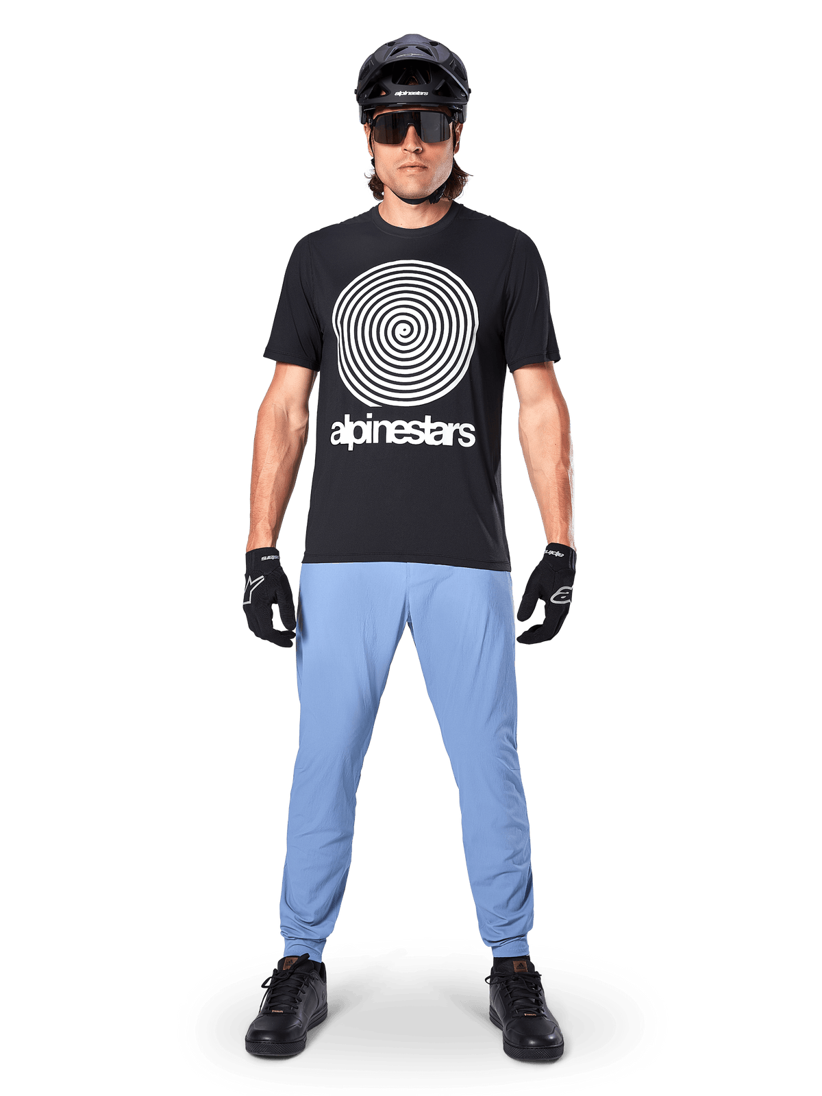 Alpinestars Maglia A-Dura Oscar a maniche corte, maglia da MTB nera con grande grafica a spirale Oscar vintage bianca e testo del brand, indossata con pantaloni azzurri, casco nero, occhiali da sole e guanti neri, abbigliamento tecnico da mountain bike