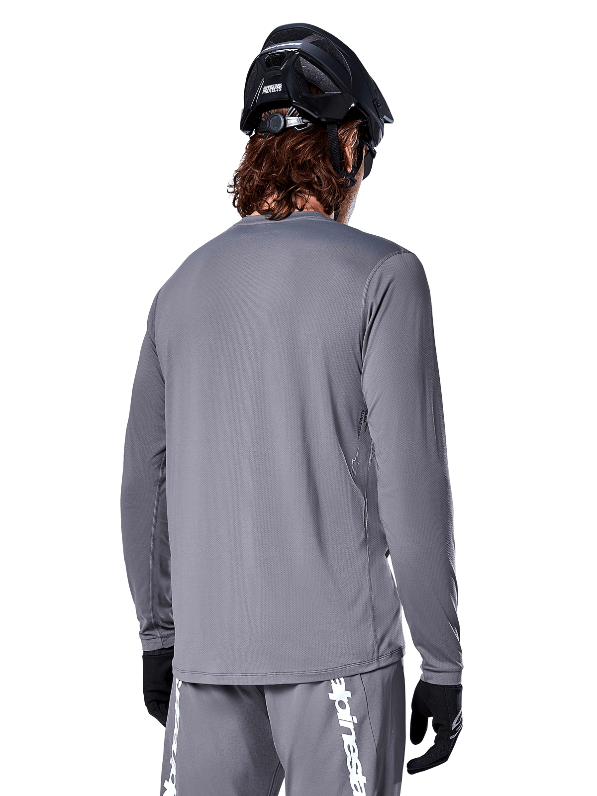 Maglia a maniche lunghe Alpinestars A-Dura Rocker in grigio scuro, vista posteriore di un ciclista di mountain bike, abbinata a pantaloncini grigi coordinati con branding bianco, casco da MTB nero e guanti neri, equipaggiamento professionale per ciclismo fuoristrada.