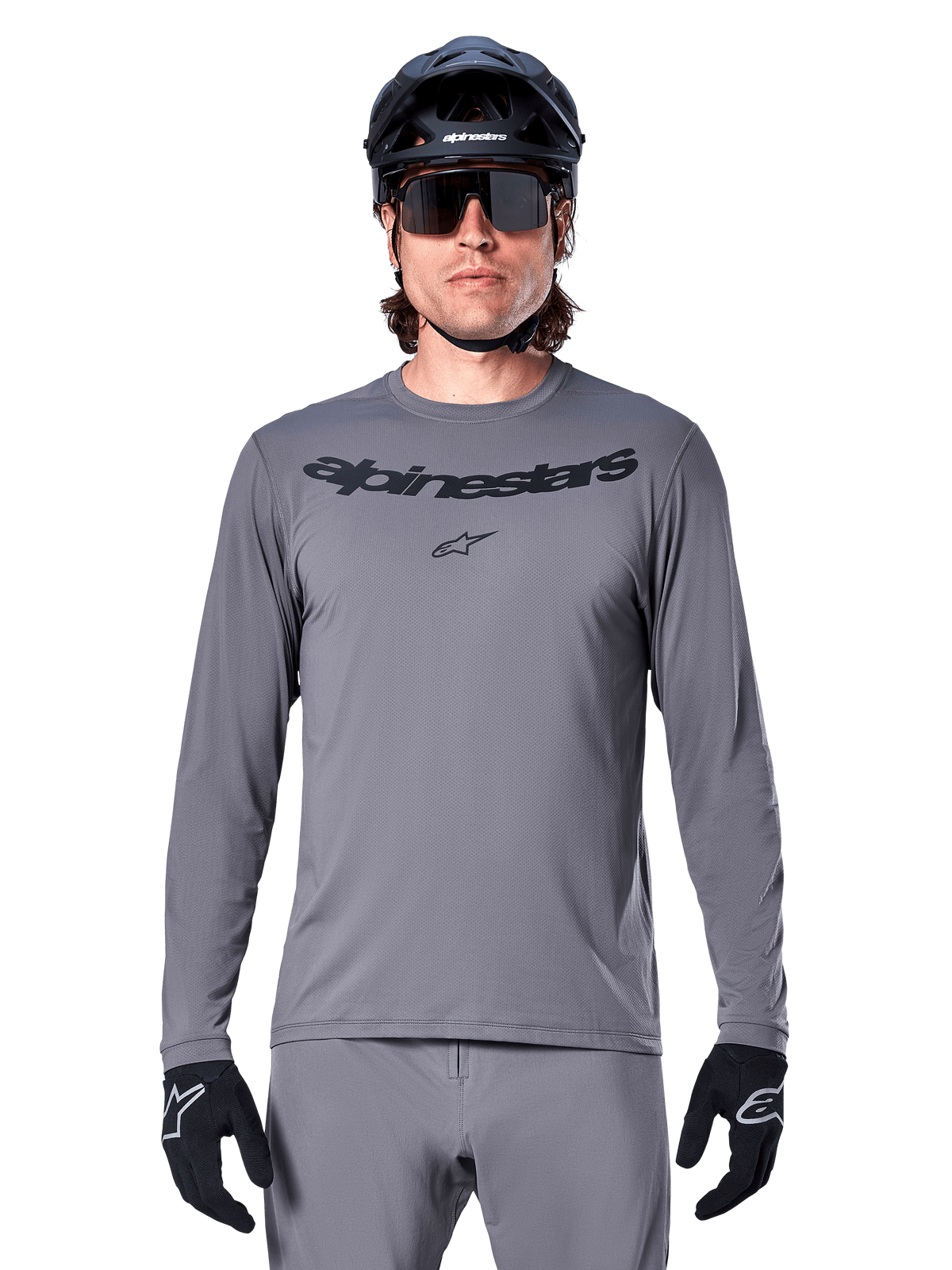 Ciclista che indossa la maglia Alpinestars A-Dura Rocker a maniche lunghe, maglia da MTB grigio scuro con logo nero sul petto, abbinata a pantaloni grigi, casco nero e guanti, progettata per il ciclismo fuoristrada