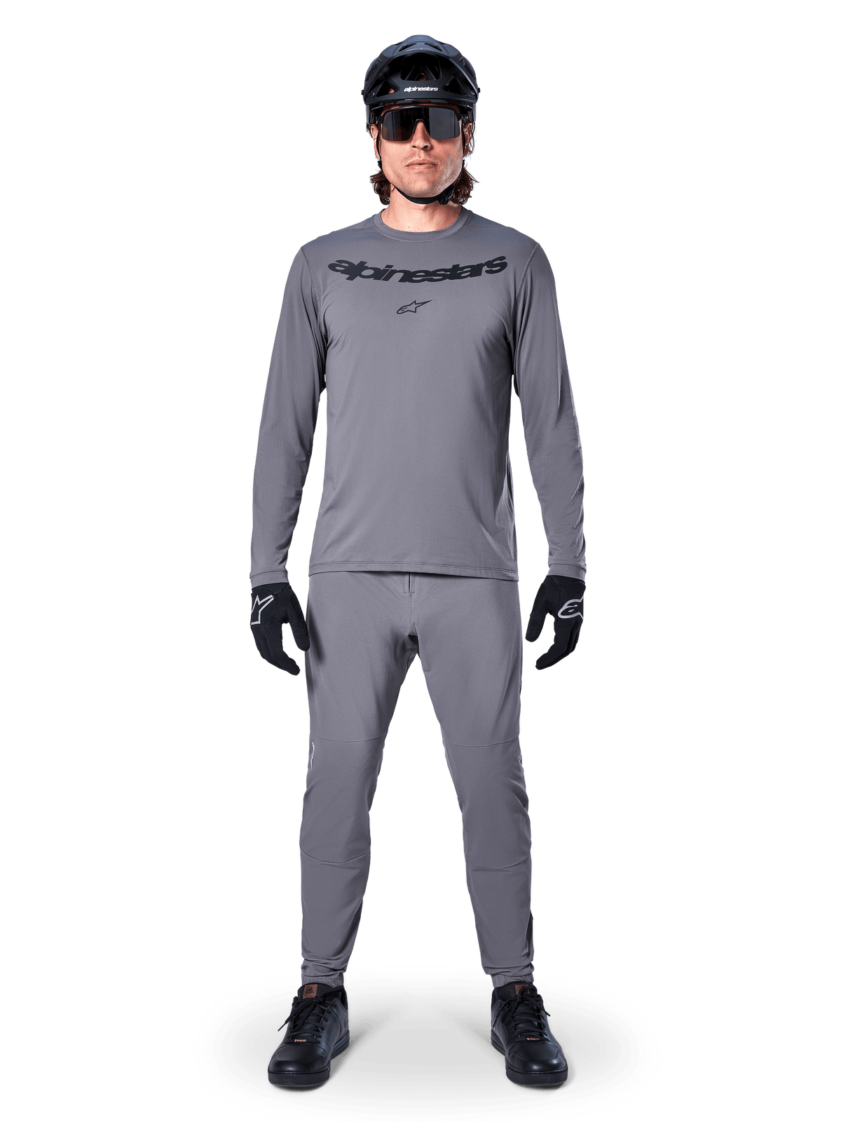 Ciclista che indossa la maglia a maniche lunghe Alpinestars A-Dura Rocker in grigio scuro, pantaloni grigi coordinati, casco da MTB nero, occhiali da sole e guanti, abbigliamento professionale da mountain bike per sentieri off-road