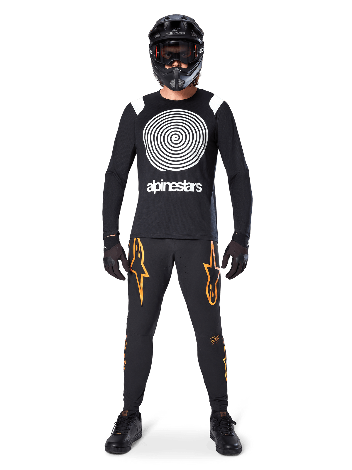 Modello con equipaggiamento completo da mountain bike, maglia a maniche lunghe Alpinestars A-Supra Oscar nera con grafica a spirale bianca sul petto, pantaloni neri con loghi a stella arancioni, casco integrale e maschera neri, guanti e scarpe neri, outfit coordinato per MTB gravity