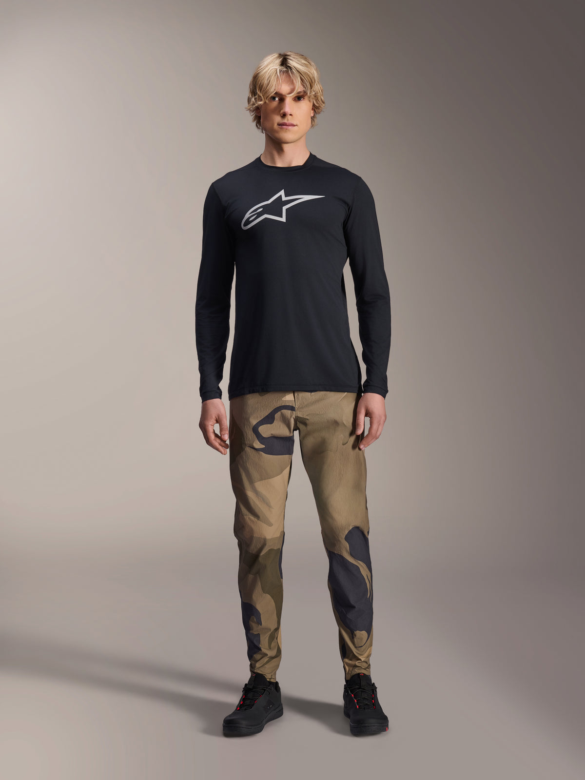 Maglia a maniche lunghe Alpinestars A-Dura Astar, nero, con un grande logo Astars bianco sul petto, indossata con pantaloni mimetici e scarpe nere, abbigliamento casual performante per mountain bike