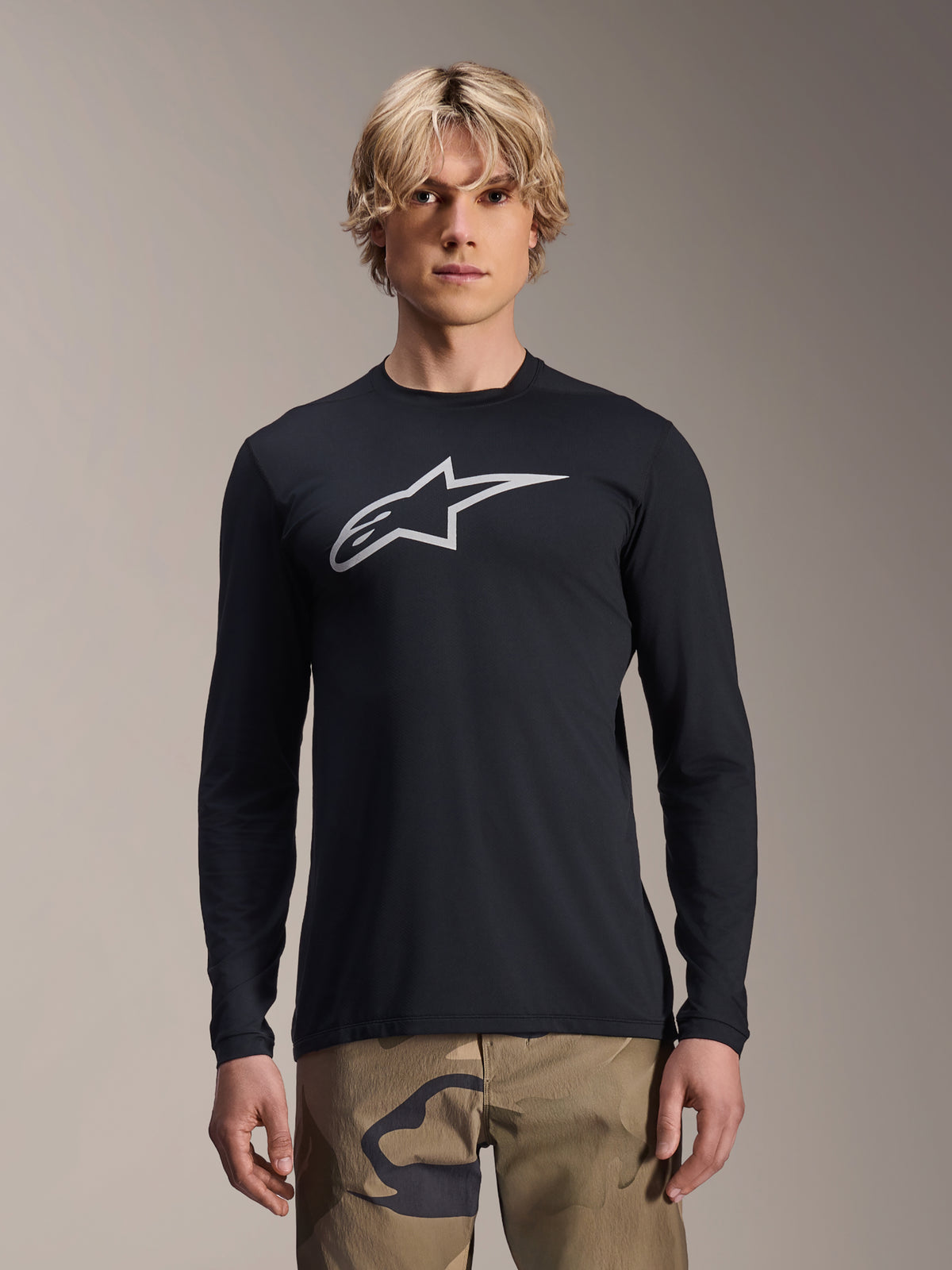 Alpinestars Maglia A-Dura Astar a maniche lunghe, nera con logo Astars argento sul petto, indossata da un modello con pantaloni mimetici, abbigliamento tecnico casual per mountain bike