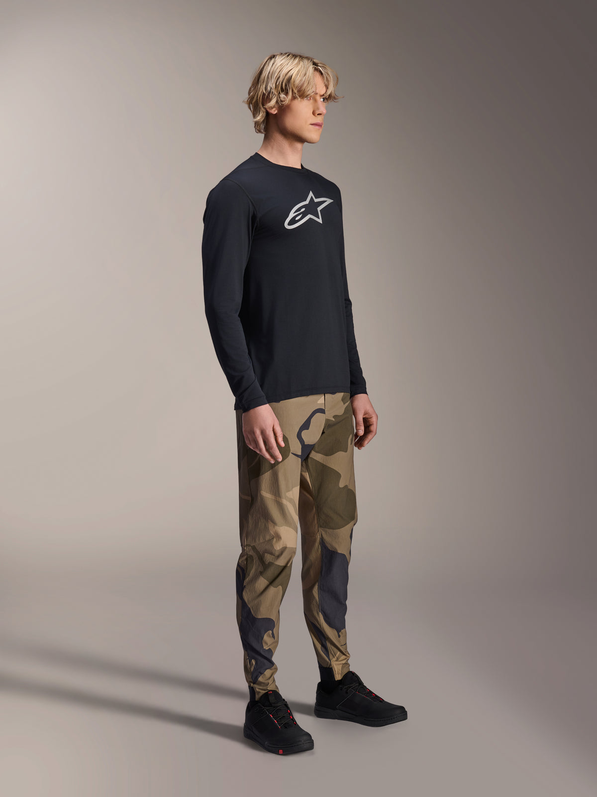 Alpinestars pantaloni A-Dura Camo, pantaloni da MTB, camouflage marrone, indossati con maglia a maniche lunghe A-Dura Astar nera e scarpe nere, abbigliamento tecnico da mountain bike per trail riding.