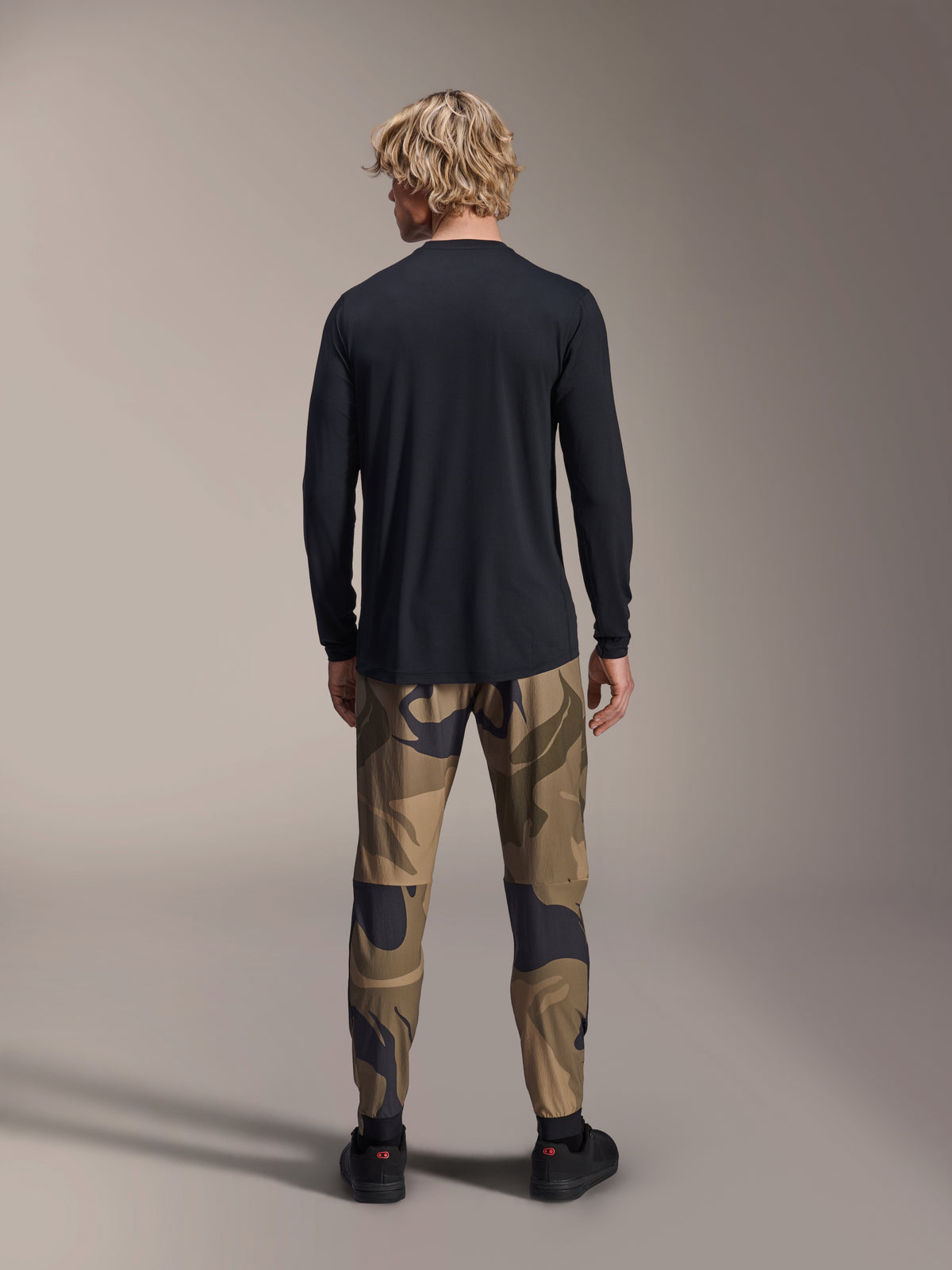 Alpinestars pantaloni A-Dura Camo, pantaloni da mountain bike, motivo mimetico marrone, vista posteriore, indossati con maglia nera a maniche lunghe e sneaker nere, pantaloni in tessuto con vestibilità regolare per ciclismo off-road