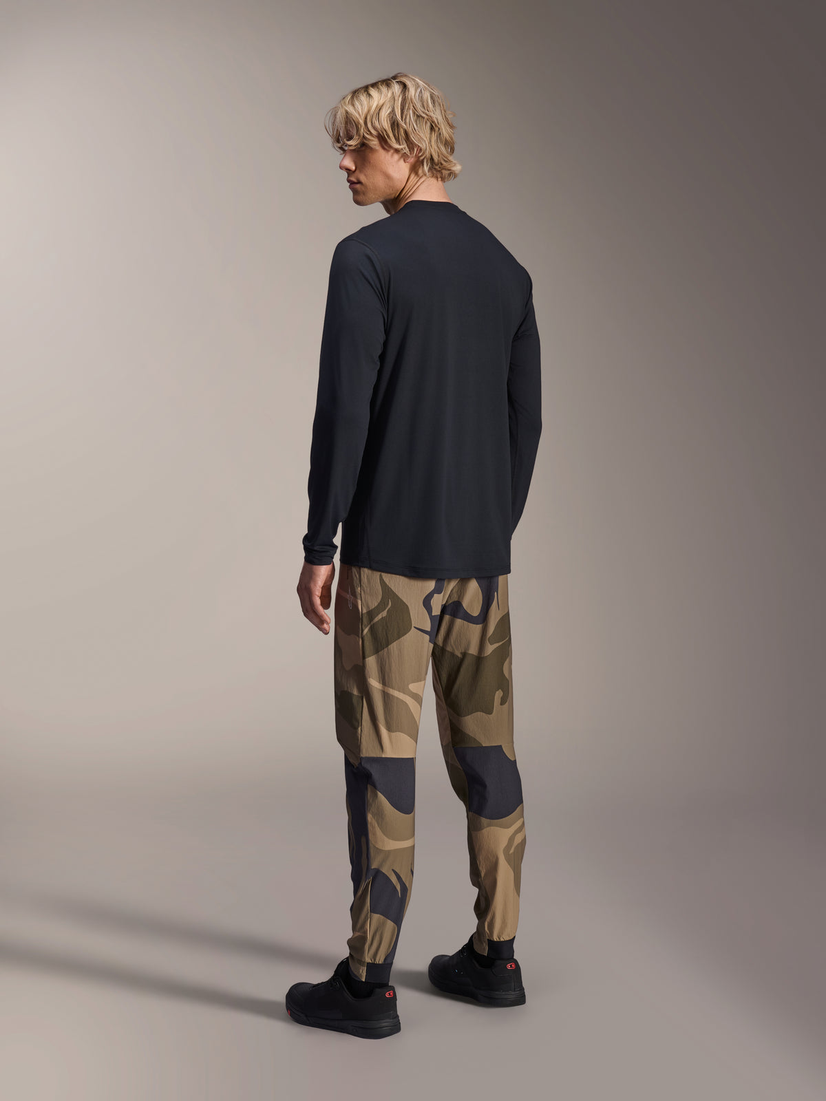 Pantaloni Alpinestars A-Dura Camo, pantaloni da MTB, mimetico marrone, indossati con maglia nera a maniche lunghe e sneaker nere, vista posteriore con gambe affusolate e polsini elasticizzati, abbigliamento tecnico per ciclismo fuoristrada