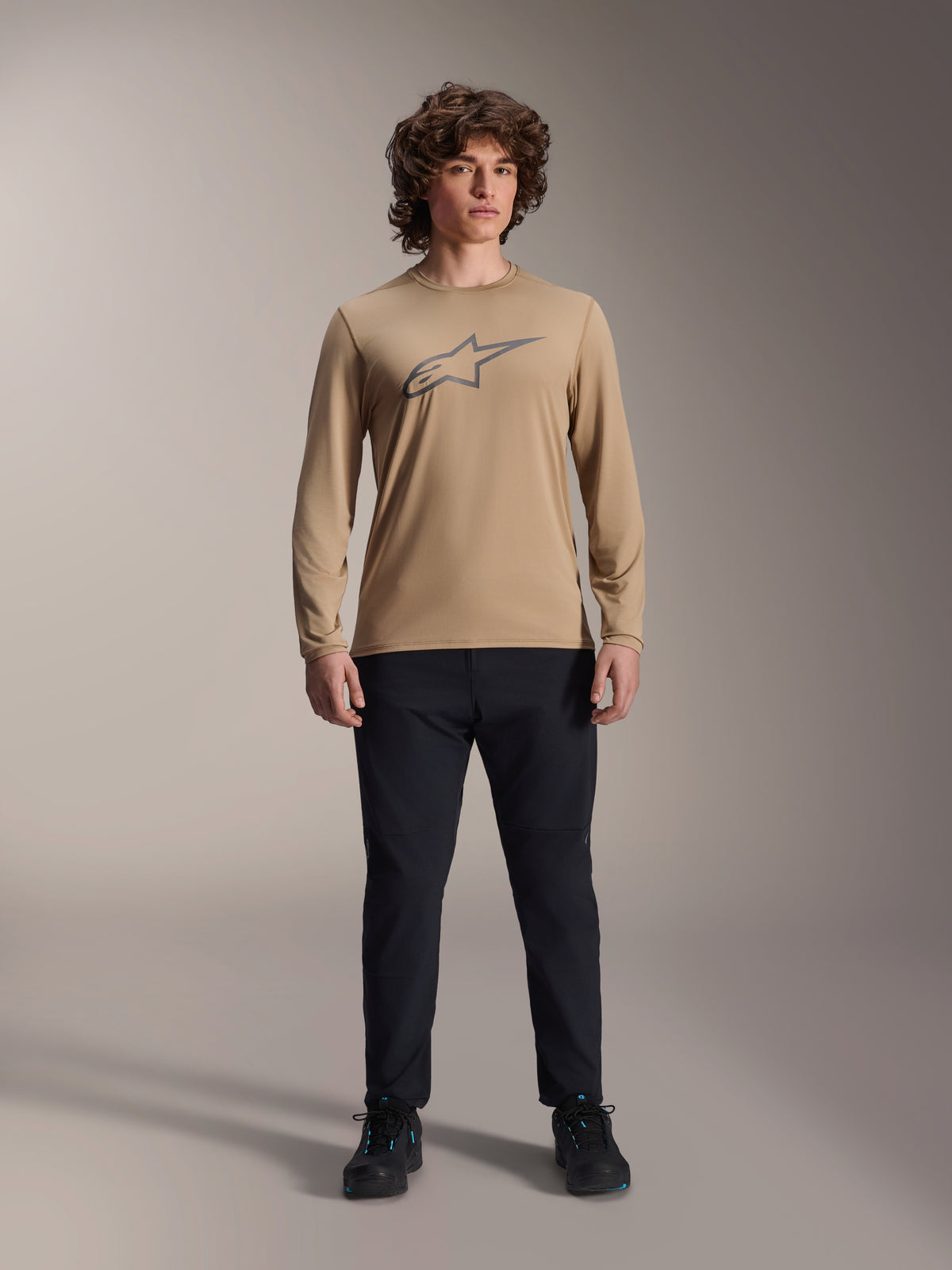 Alpinestars A-Dura Astar Long Sleeve Jersey, maglia a maniche lunghe marrone chiaro con logo Astars grigio sul petto, indossata da un modello con pantaloni tecnici e scarpe neri, abbigliamento tecnico casual per mountain bike