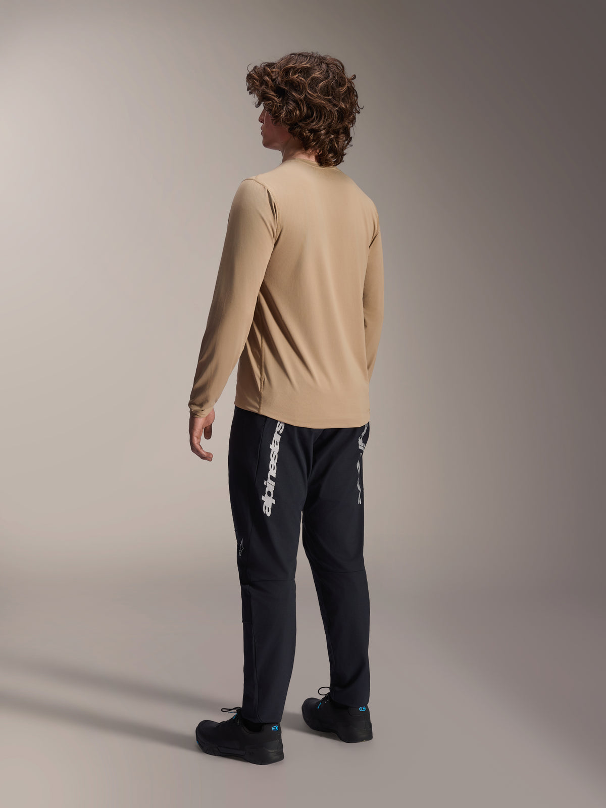 Maglia a maniche lunghe Alpinestars A-Dura Astar in marrone chiaro, vista posteriore di un modello maschile che mostra il design a coda allungata, abbinata a pantaloni e scarpe neri, abbigliamento tecnico casual per mountain bike.