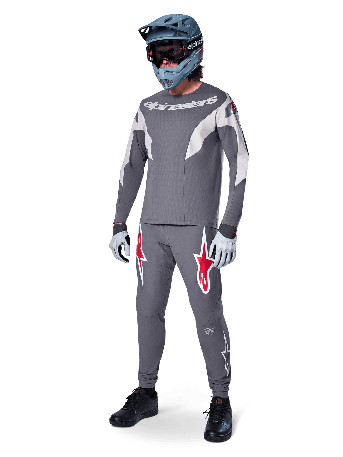 Pilota di MTB con maglia Alpinestars A-Supra Race a maniche lunghe, grigio scuro con dettagli grigio chiaro, pantaloni coordinati con logo a stella rosso, casco integrale grigio con maschera e guanti, completo tecnico per gare di mountain bike