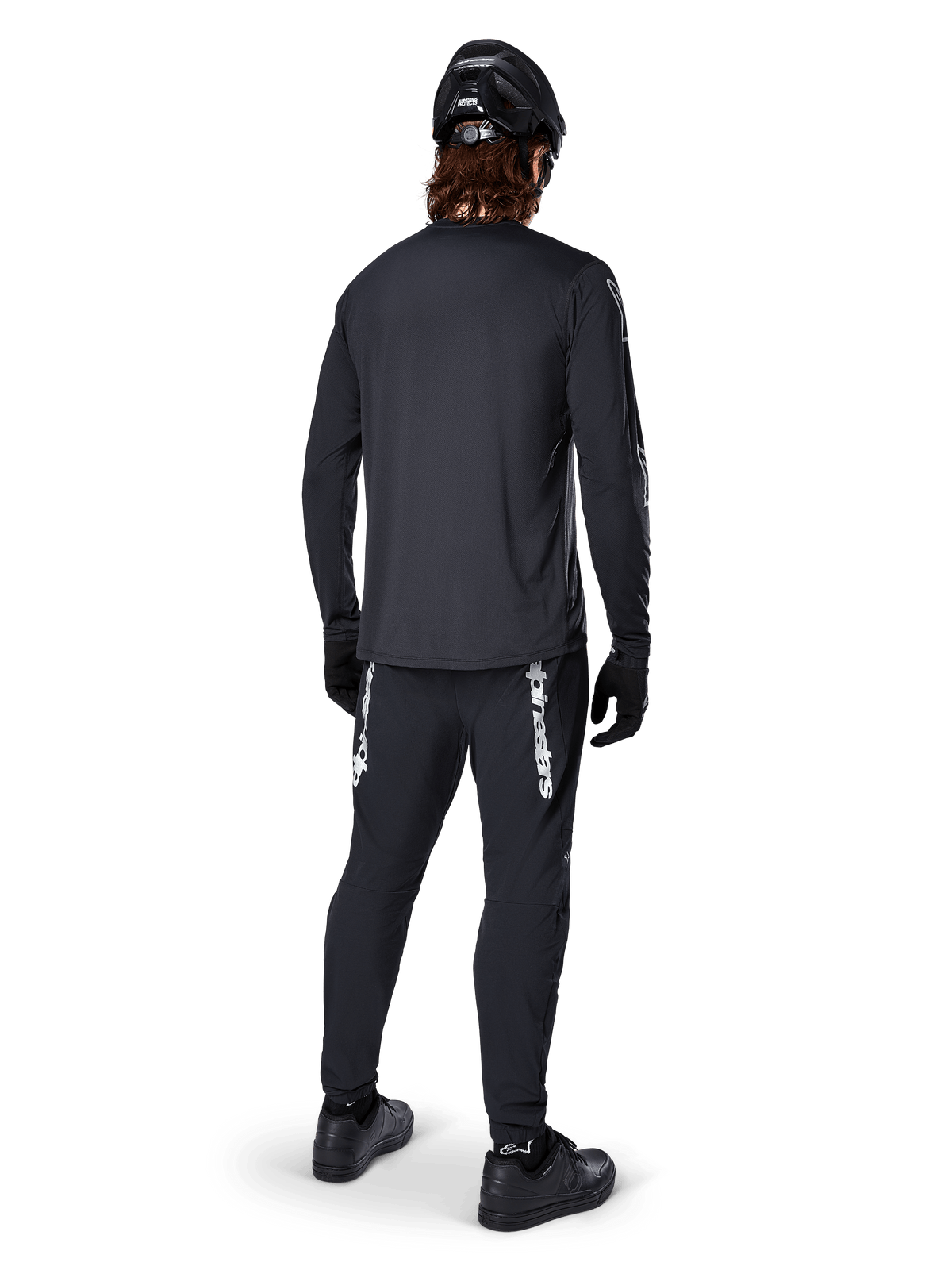 Maglia a maniche lunghe Alpinestars A-Dura Switch, nero, vista posteriore su un ciclista, indossata con pantaloni neri con logo Alpinestars bianco, casco nero e guanti, dotata di coda allungata per maggiore copertura, abbigliamento tecnico da mountain bike