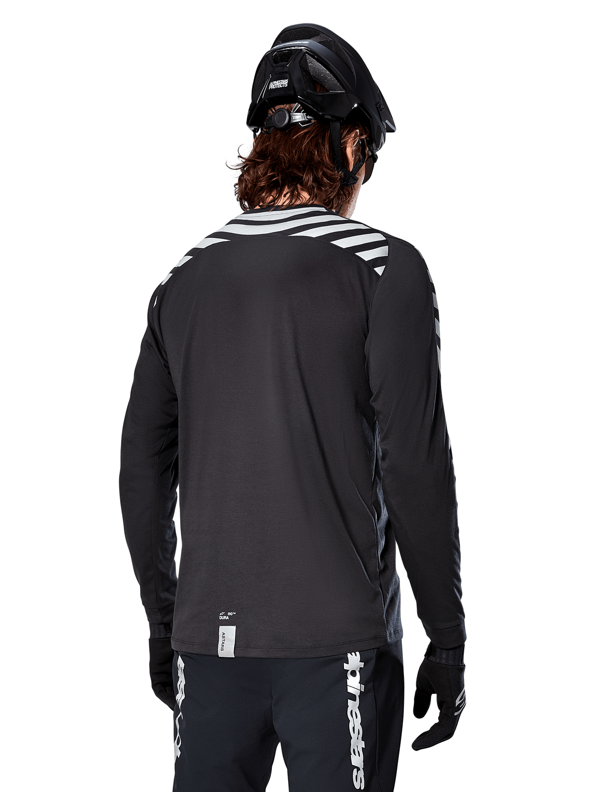 Maglia a maniche lunghe Alpinestars A-Dura Dri Oscar, Nero Riflettente, vista posteriore con strisce diagonali riflettenti sulle spalle e una coda più lunga, indossata con casco e guanti da MTB neri, equipaggiamento tecnico per mountain bike
