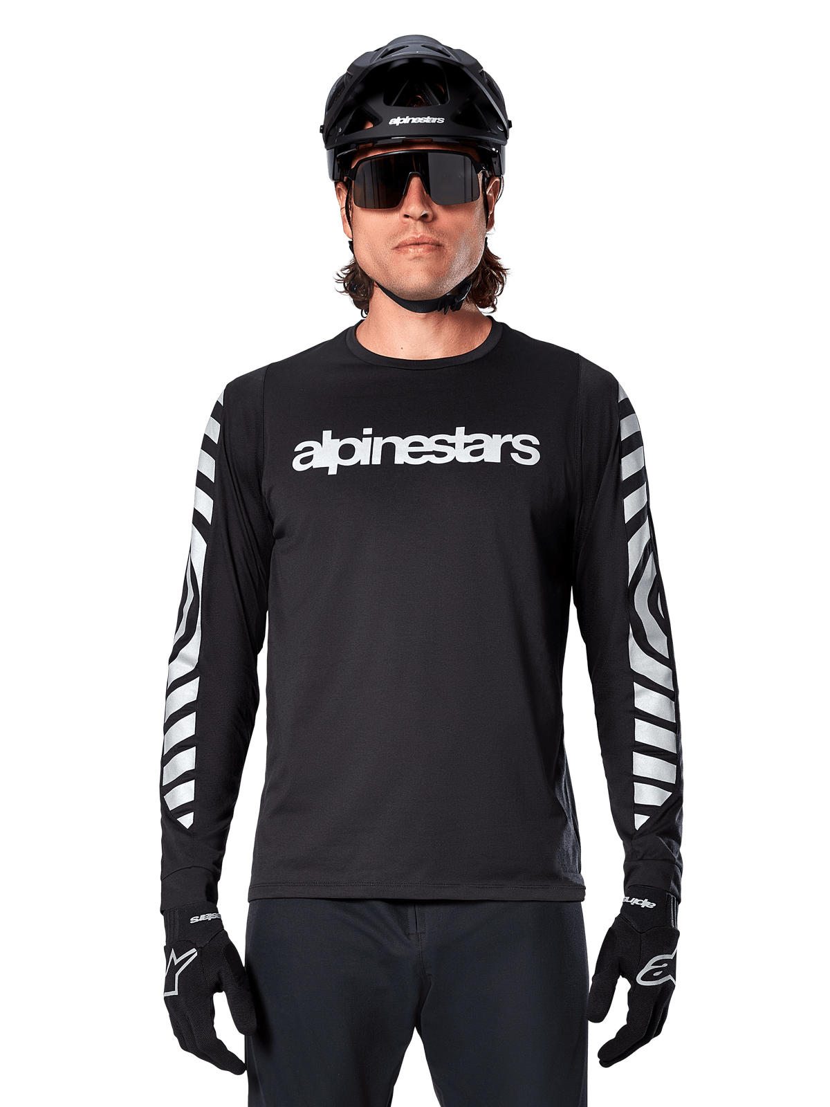 Ciclista uomo che indossa la maglia a maniche lunghe Alpinestars A-Dura Dri Oscar, Black Reflective, con logo Alpinestars bianco sul petto e grafiche riflettenti argento sulle maniche, abbinata a casco da MTB nero, occhiali da sole e guanti neri, abbigliamento tecnico da ciclismo