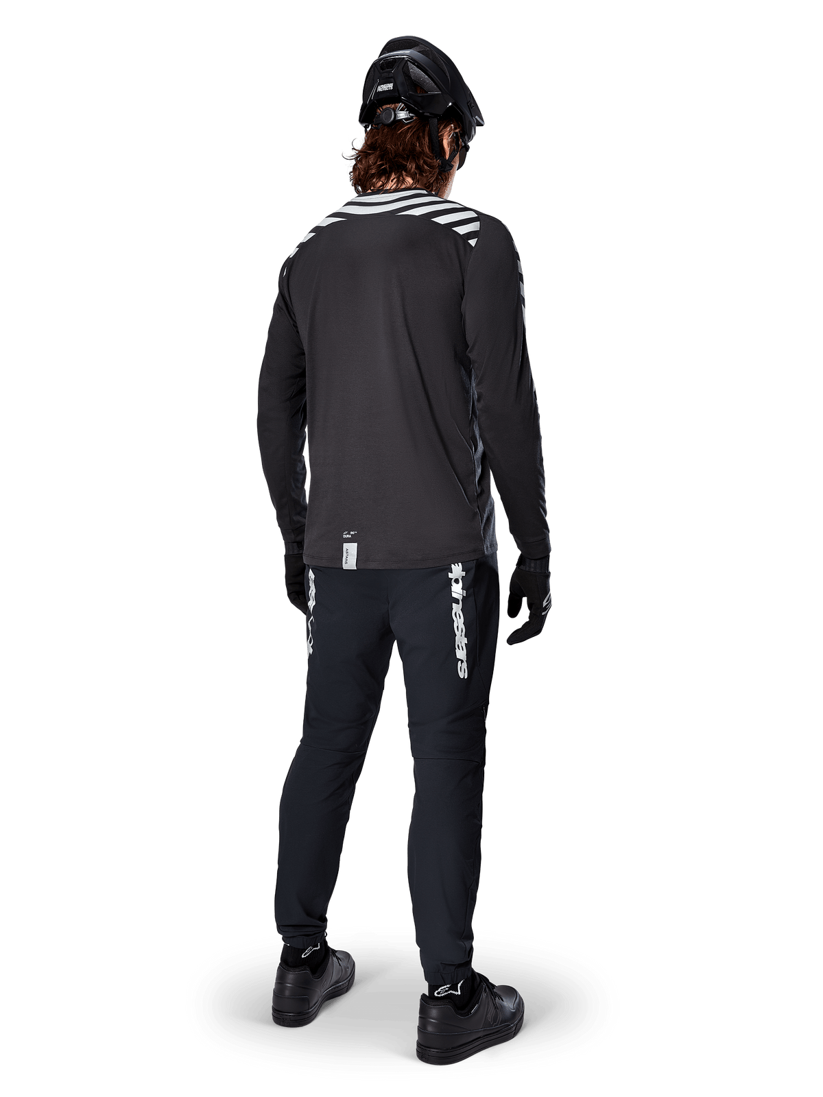 Ciclista di mountain bike con maglia a maniche lunghe Alpinestars A-Dura Dri Oscar in Black Reflective, vista da dietro, abbinata a pantaloni tecnici neri, casco e guanti, con grafiche a strisce riflettenti sulle spalle e coda posteriore allungata per il ciclismo fuoristrada.