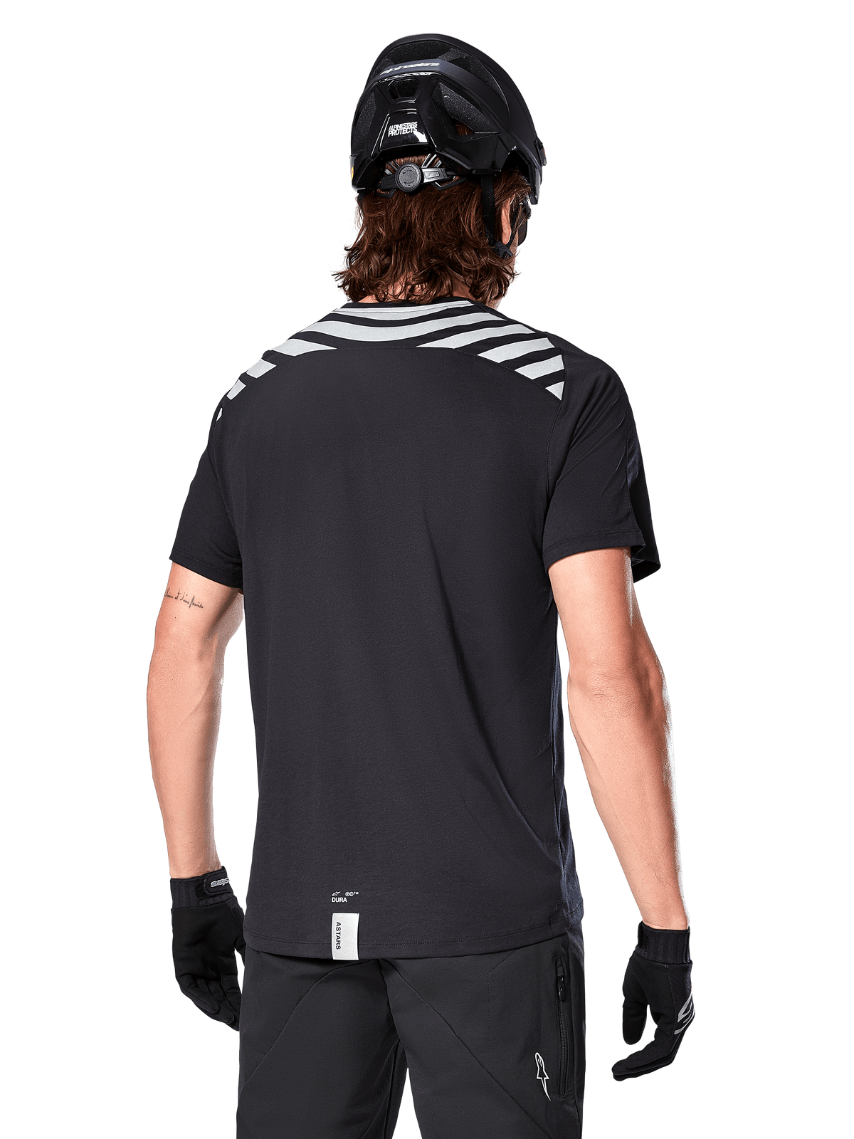 Maglia Alpinestars A-Dura Dri Oscar, maglia da MTB a maniche corte, Nero Riflettente, vista posteriore su modello con casco e guanti da ciclismo, con strisce riflettenti grigie sulle spalle e coda più lunga, tessuto tecnico traspirante per mountain bike